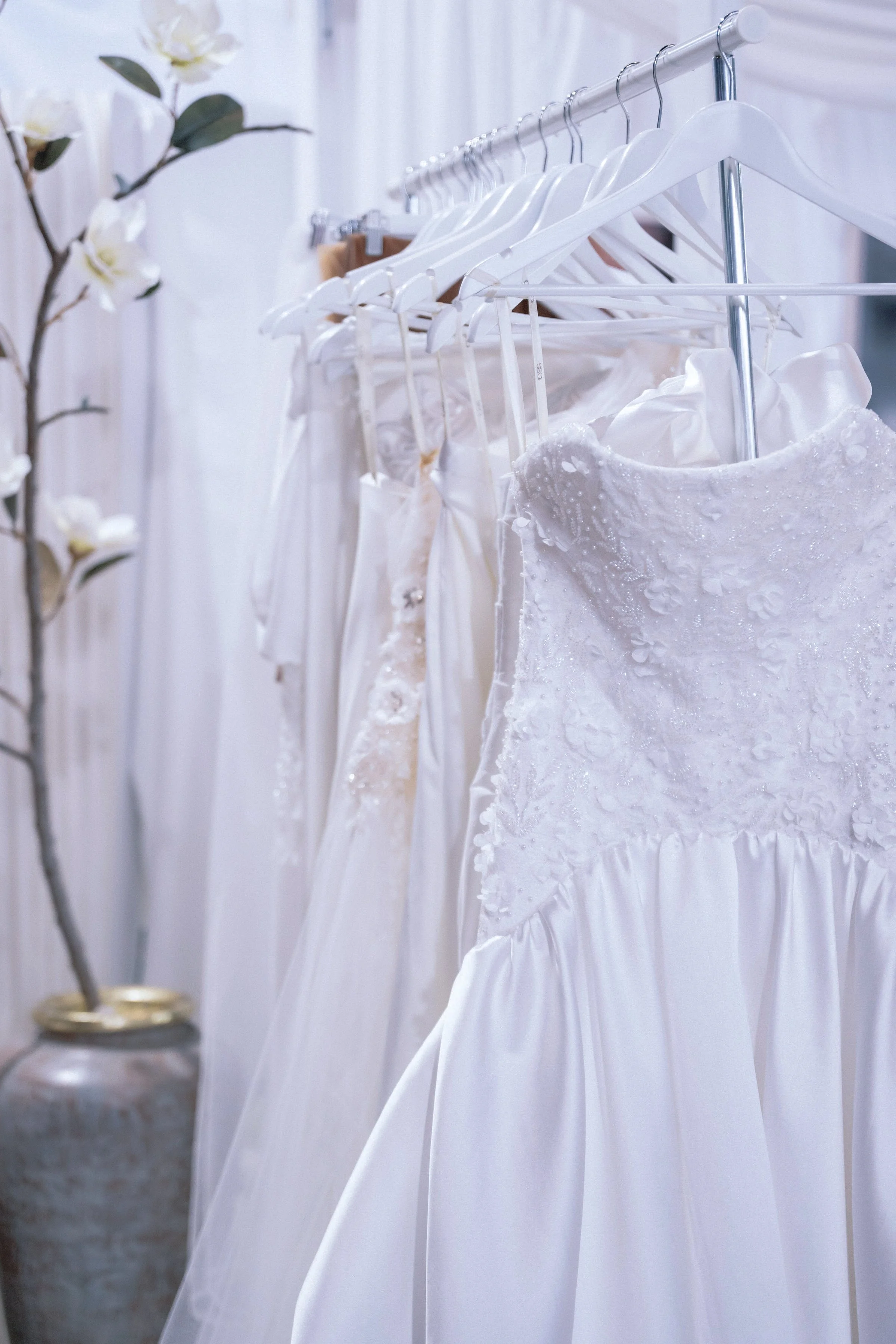 Close-up of white wedding dresses hanging on a rack, featuring floral textures and satin skirts in a soft, elegant bridal boutique setting.