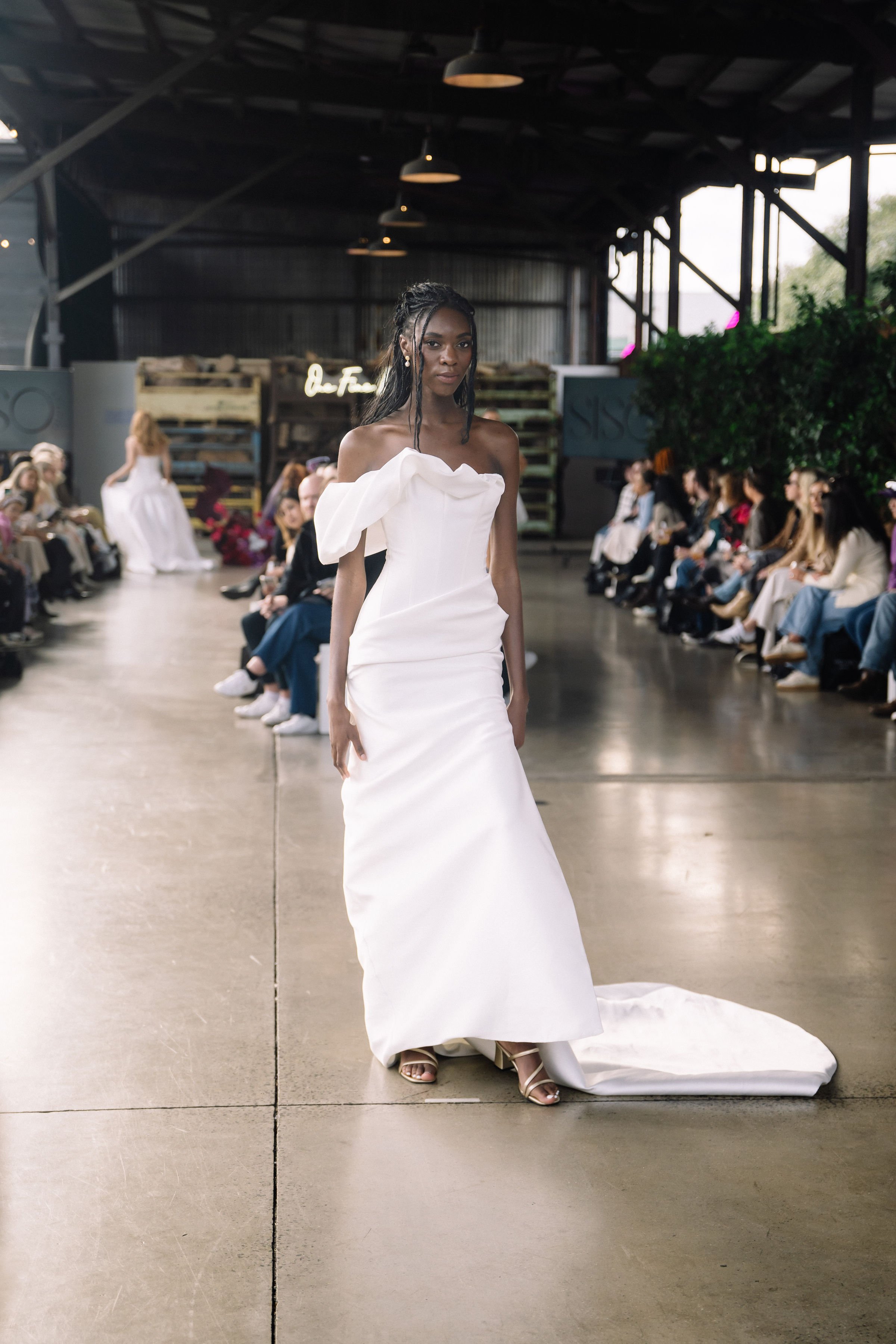 Model in a white off-shoulder gown with asymmetrical ruffle detail walks down the runway at an indoor fashion show.