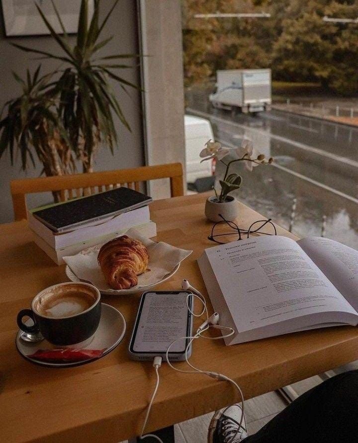 A wooden table with a cup of coffee, a croissant on a plate, a smartphone with earphones, a stack of books, an open book or notebook, glasses, and a potted orchid plant, with a window showing a rainy street scene outside.