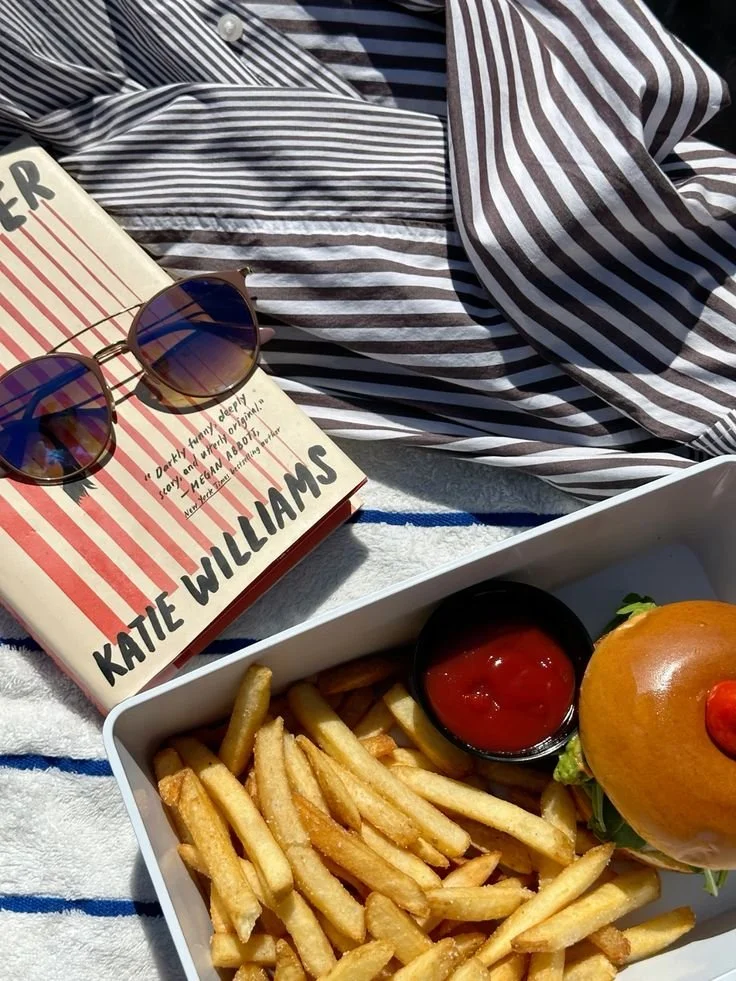 A tray of French fries with a small cup of ketchup and a cheeseburger with lettuce and tomato, on a striped cloth, accompanied by a book titled 'Fahrenheit 451' and a pair of sunglasses.