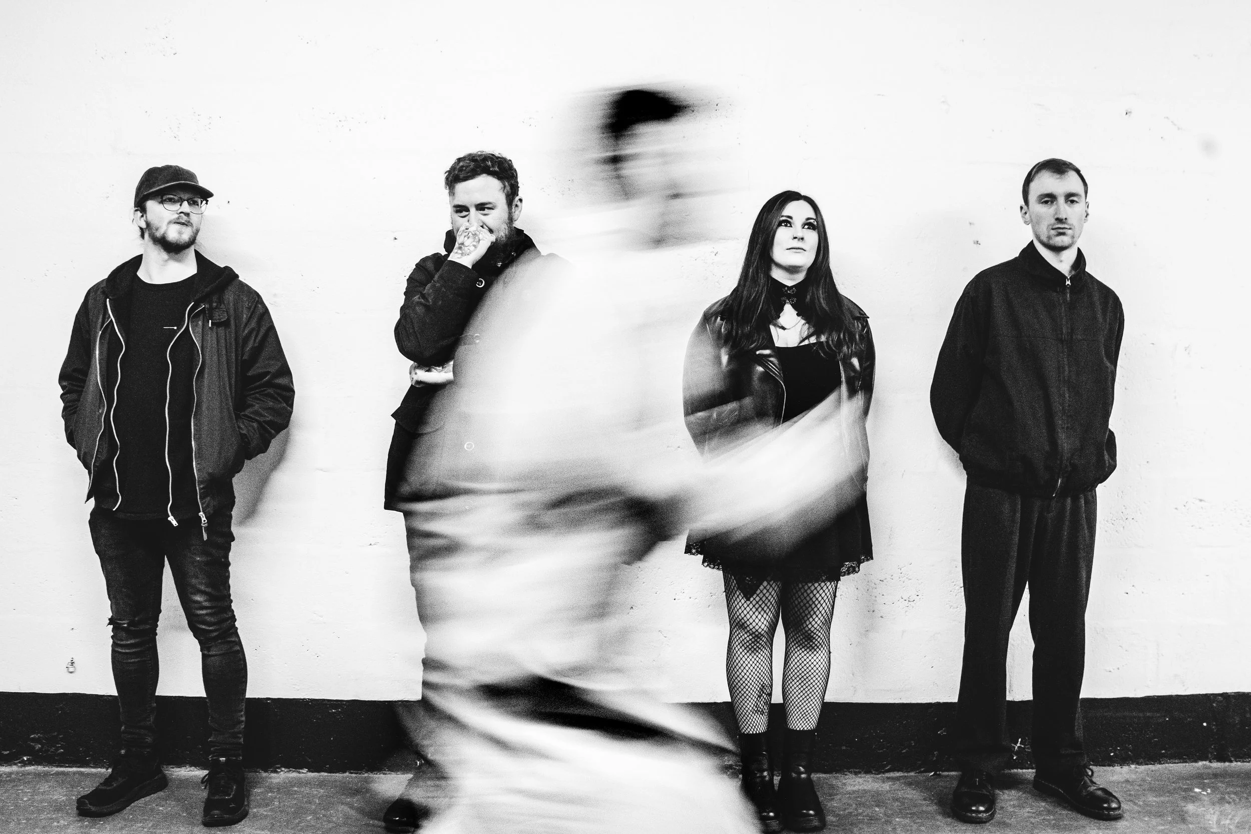 Black and white photo of five people standing against a plain wall. The second person appears to be moving, creating a blurred effect.