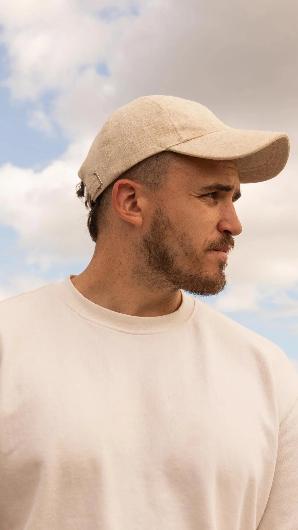 Side profile of a man with facial hair wearing a beige baseball cap and light-colored sweatshirt, standing outdoors with a cloudy sky in the background.