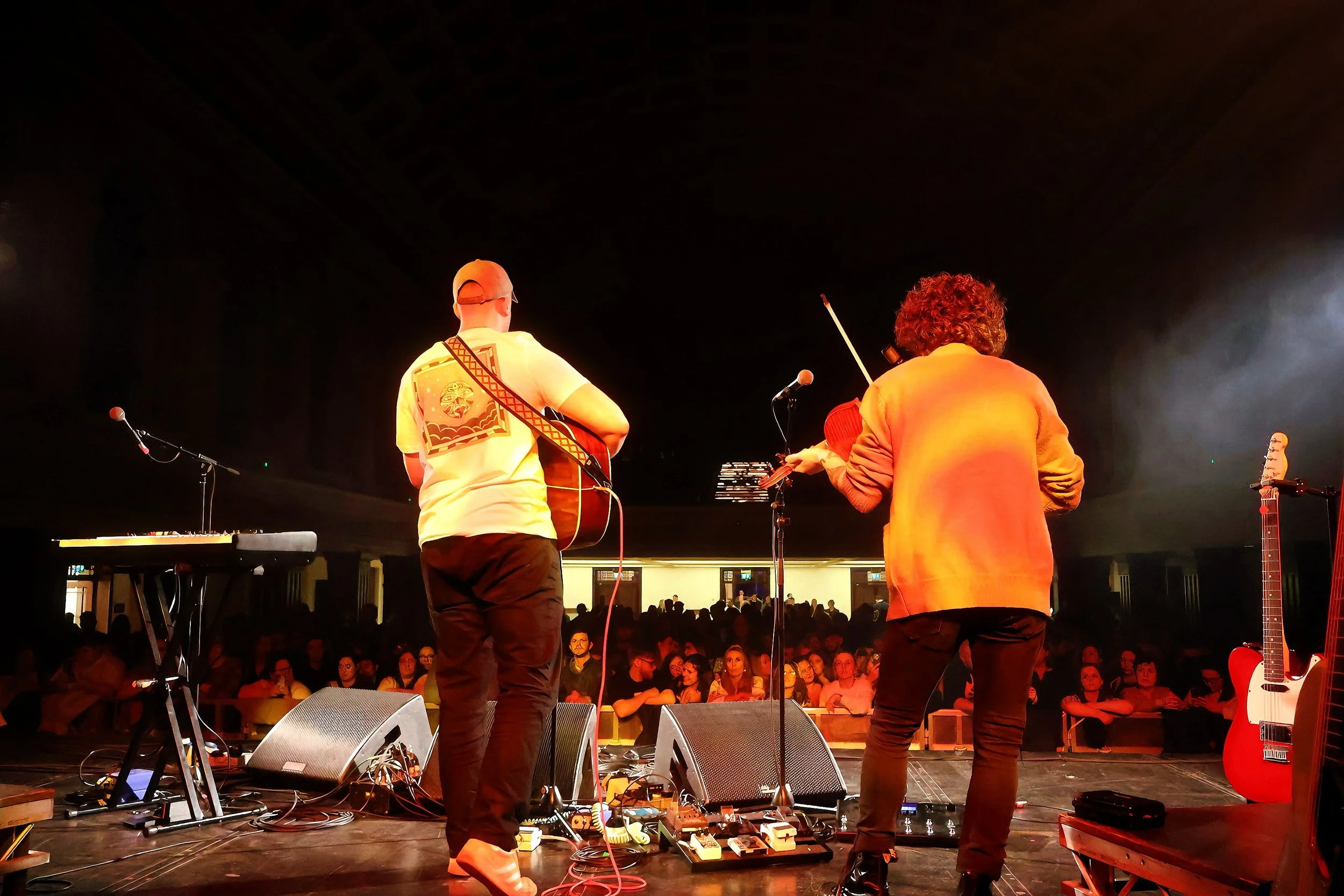 Two musicians on stage performing with guitars and a violin in front of an audience at a concert.