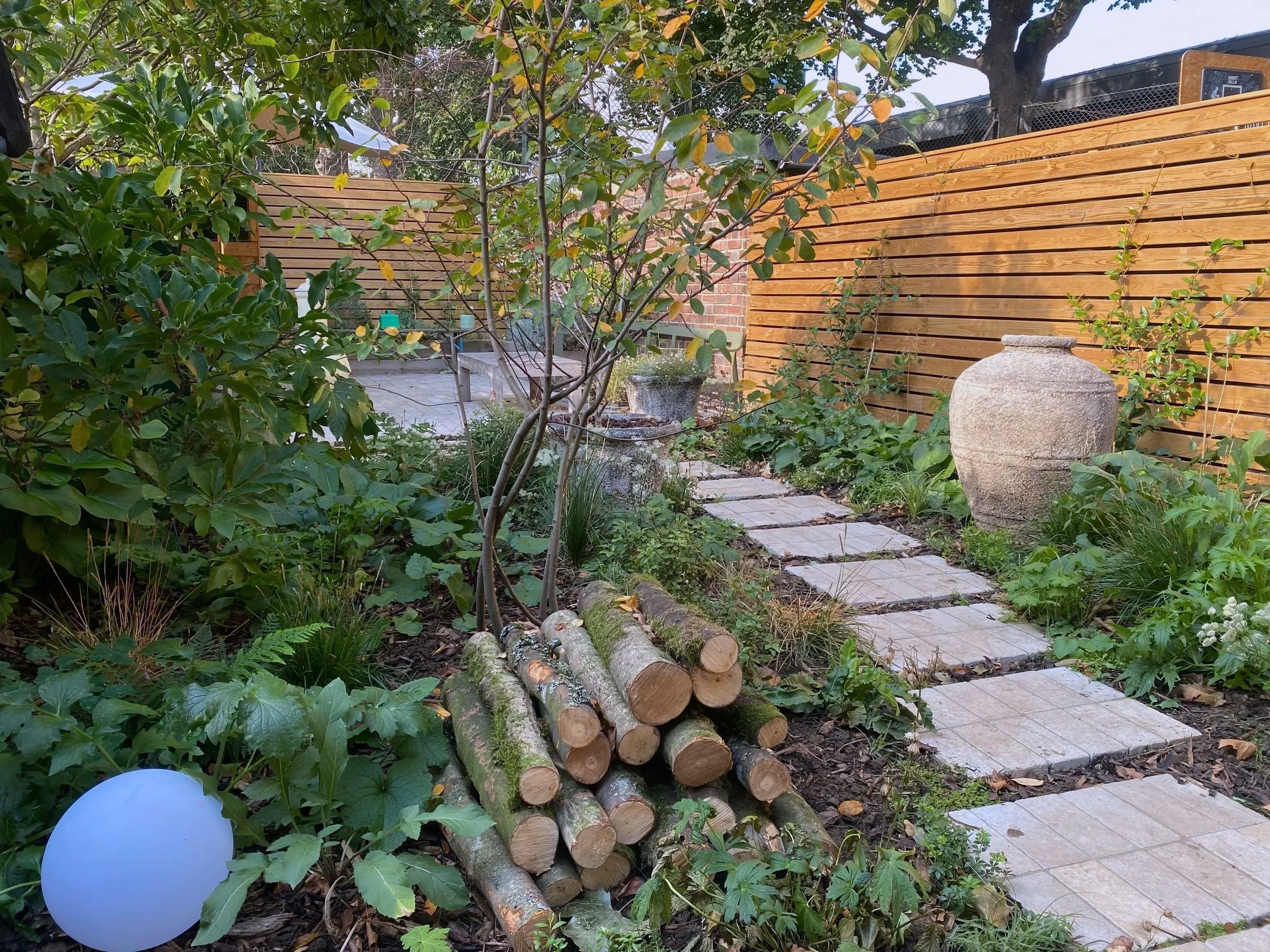 A rear London garden with a slatted fence, mixed naturalistic planting, a large stone vase and logpile. This garden has been designed by Studio Kamio, who provide high quality garden design and planting design in London.