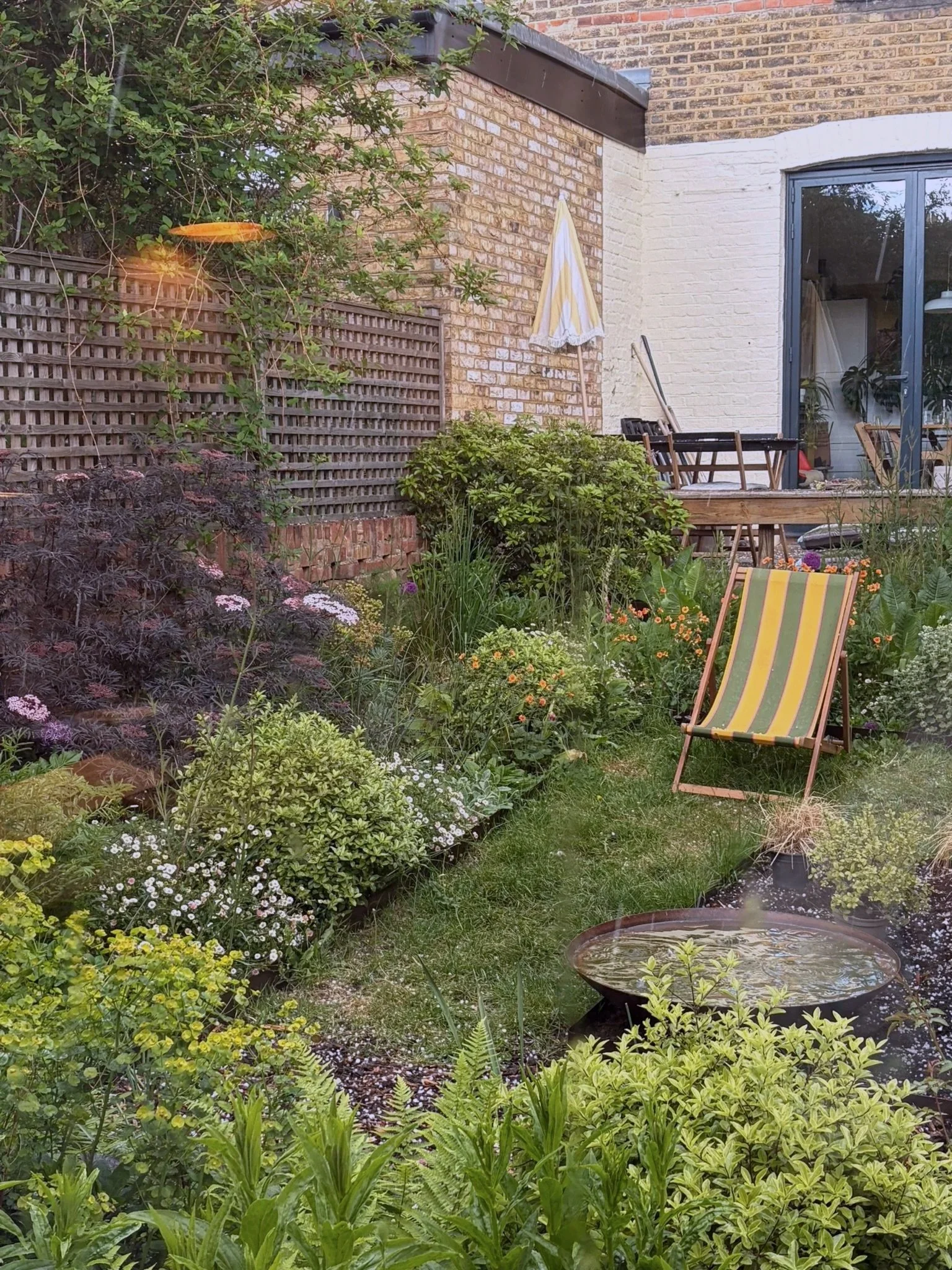 A lush backyard garden with various plants, flowers, and shrubs, a small pond, a deck with chairs, and an umbrella. This garden has been designed by Studio Kamio, who provide high quality garden design and planting design in London.