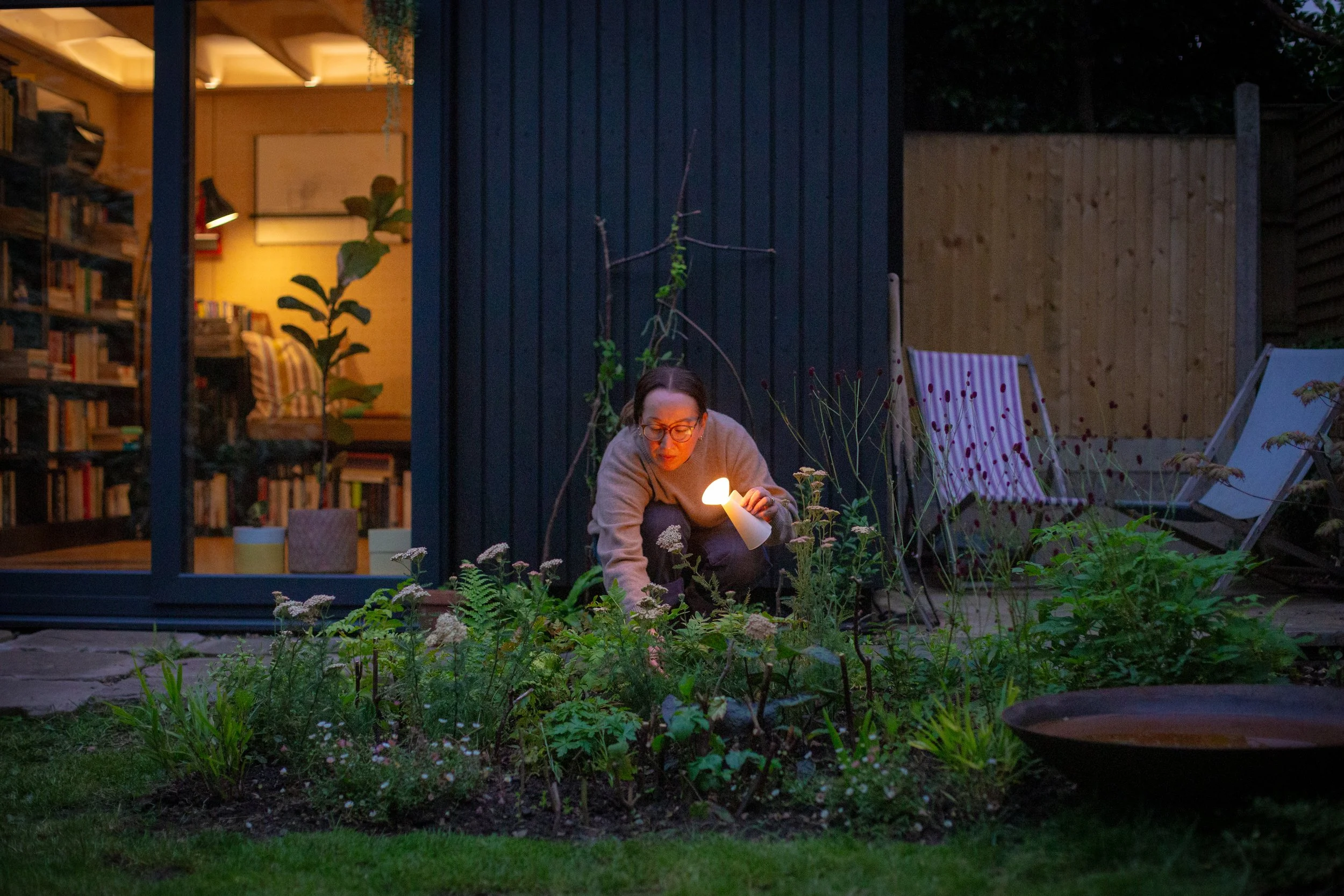 A person with glasses and long hair tending to plants in a garden during dusk, holding a small lamp. This garden has been designed by Studio Kamio, who provide high quality garden design and planting design in London.