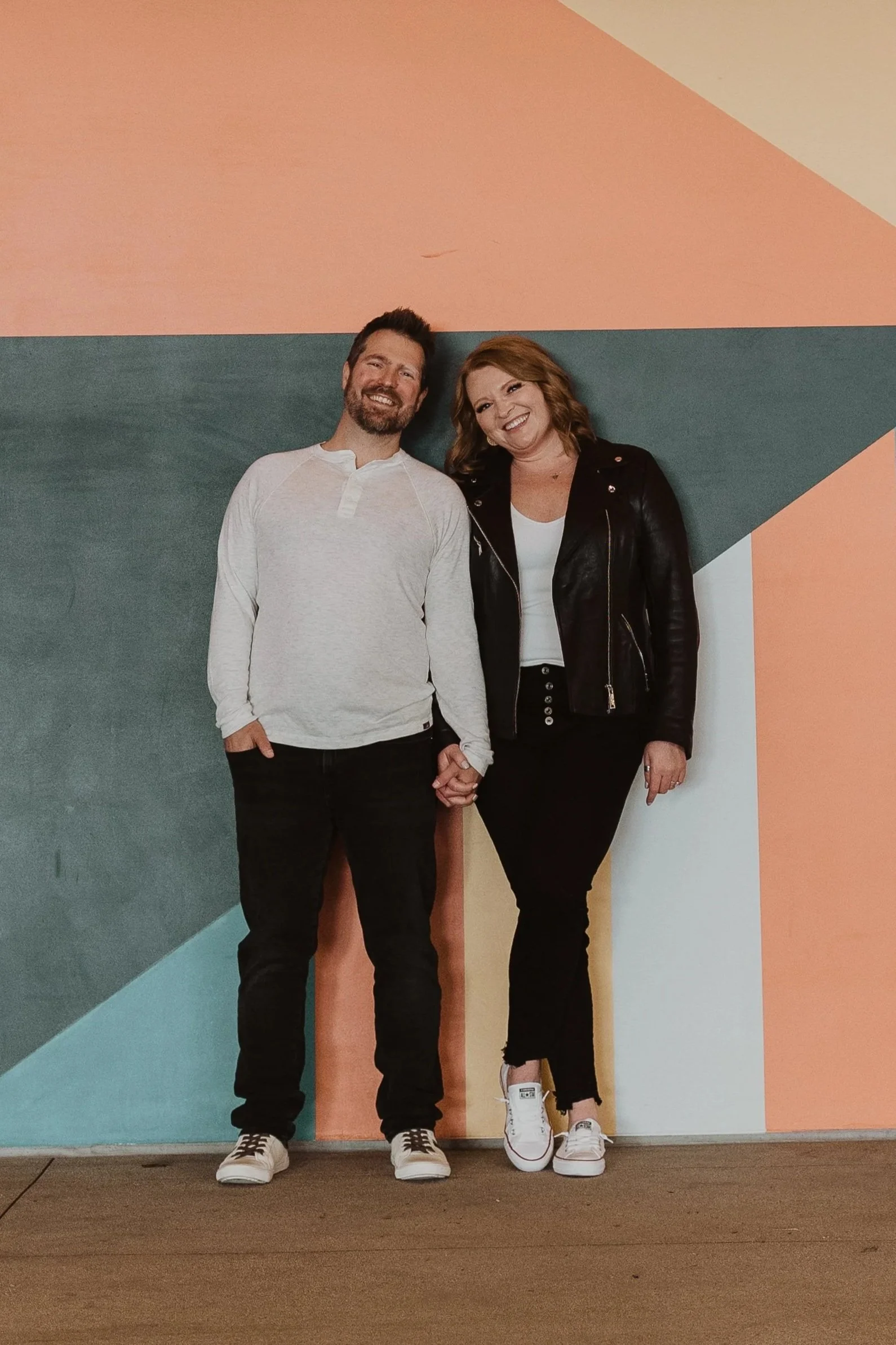 A smiling man and woman standing against a colorful geometric wall, holding hands and wearing casual outfits.