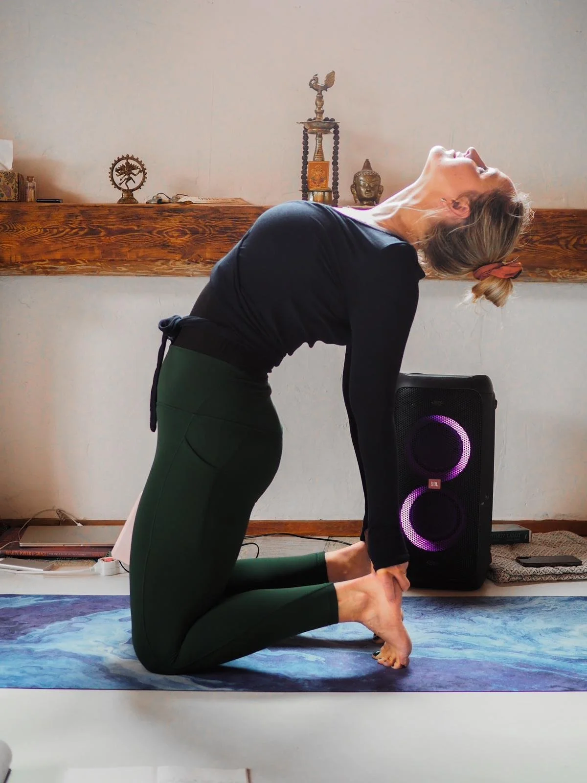 A woman practicing yoga on a mat, performing a backbend pose with her head thrown back and hands holding her heels, in a room with a wooden shelf and decorative statues in the background.