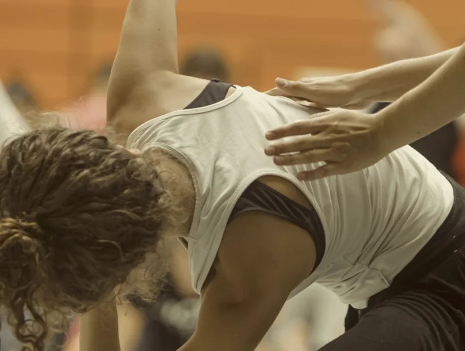 Two people practicing yoga or a similar activity, with one person bending over and the other guiding or supporting, in an indoor setting.