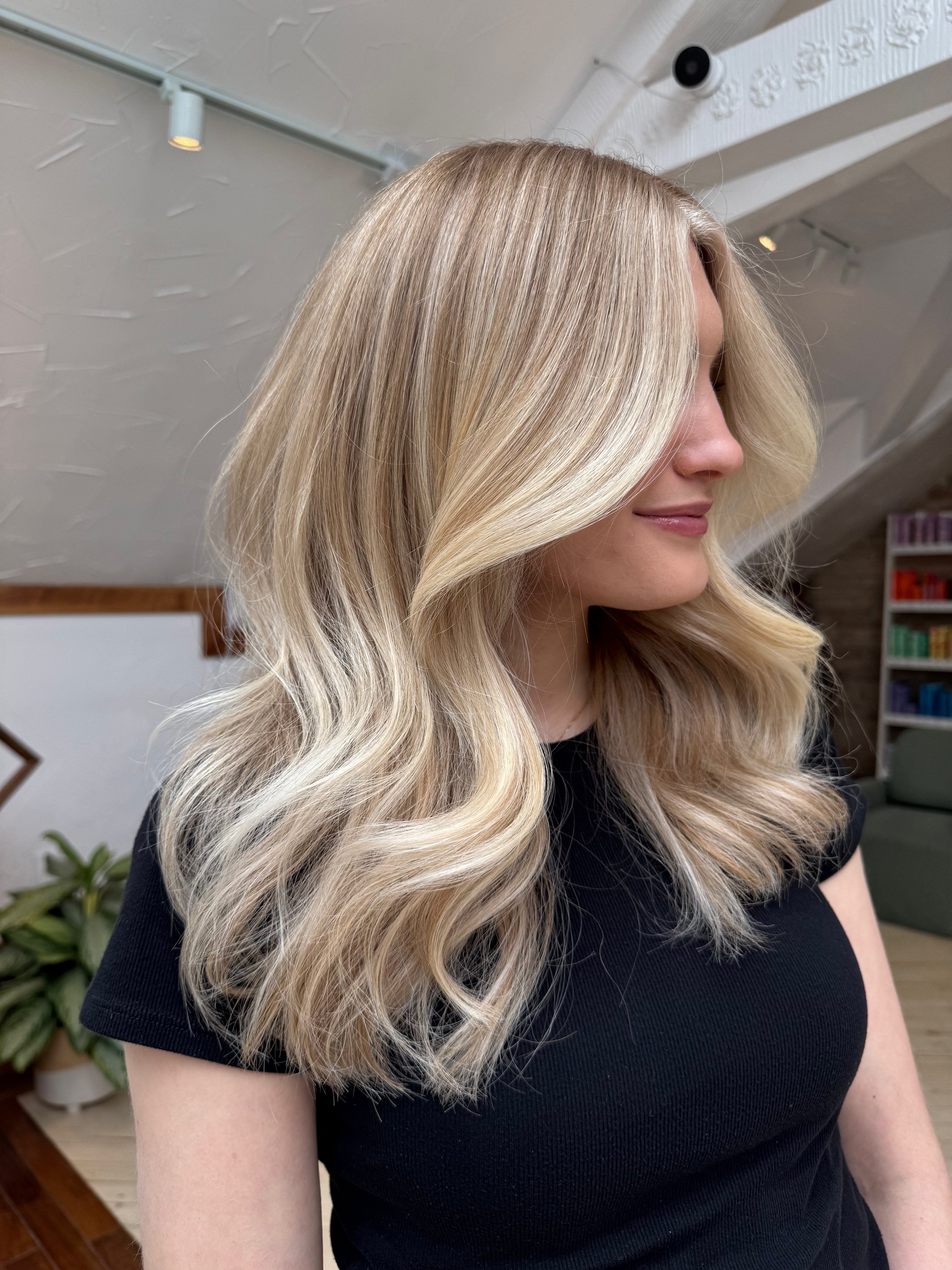 Woman with long, wavy blonde hair, wearing a black top, standing indoors with bookshelves and plants in the background.