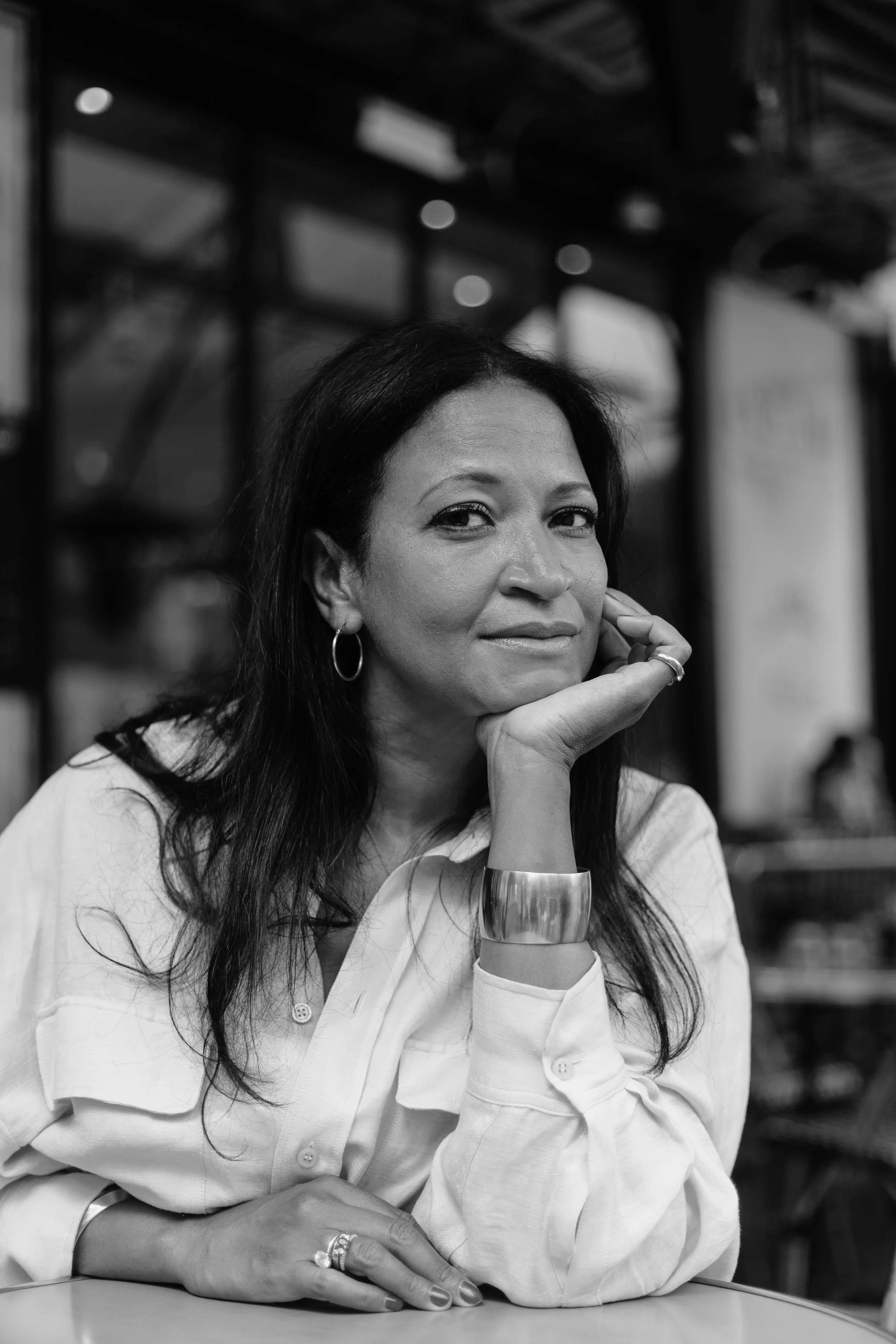A woman with dark hair and earrings, wearing a light-colored shirt, sitting at a table with her chin resting on her hand, looking at the camera in a black and white photo.