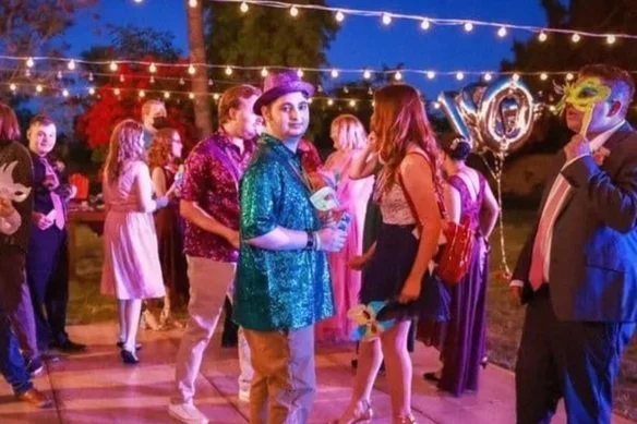Group of people celebrating at an outdoor party or dance event, with string lights overhead and some holding balloons or masks, during evening.