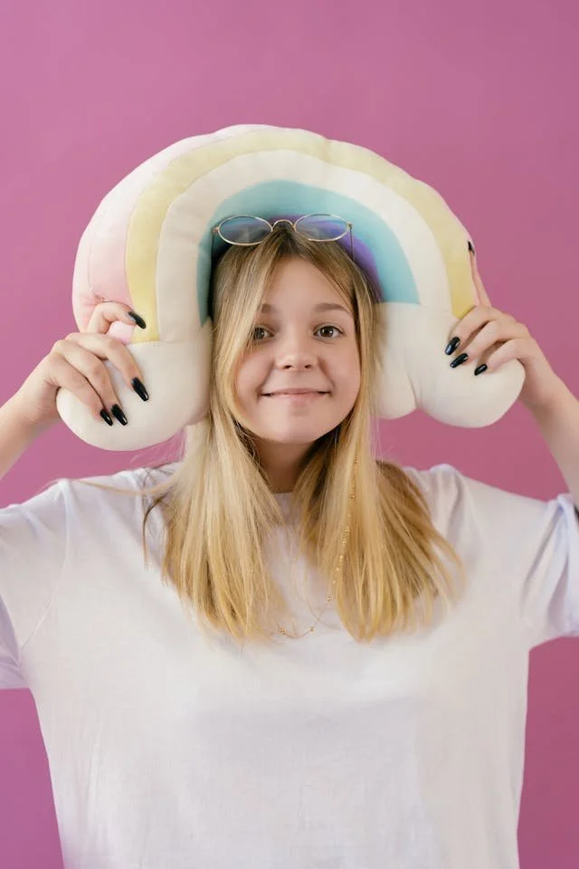 A young girl with long blonde hair smiling, wearing a white shirt, holding a rainbow-shaped pillow with a pink background.