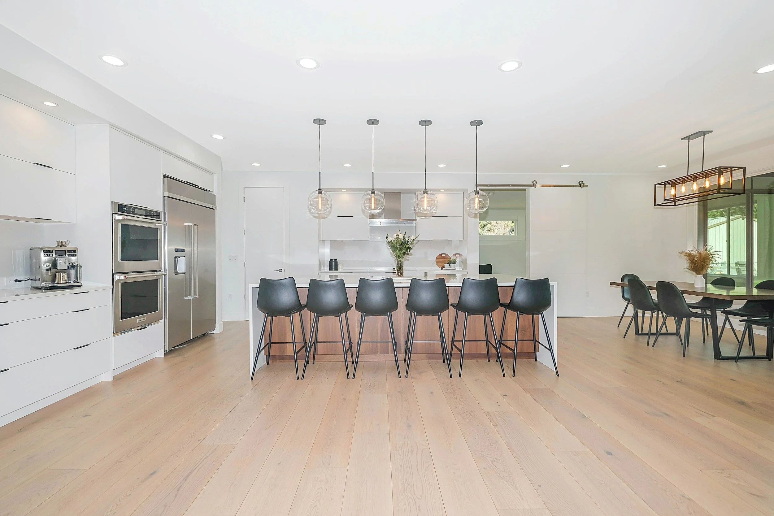 Modern open-concept kitchen with a large island, black barstools, and pendant lighting, adjacent to a dining area with black chairs and a rectangular chandelier.