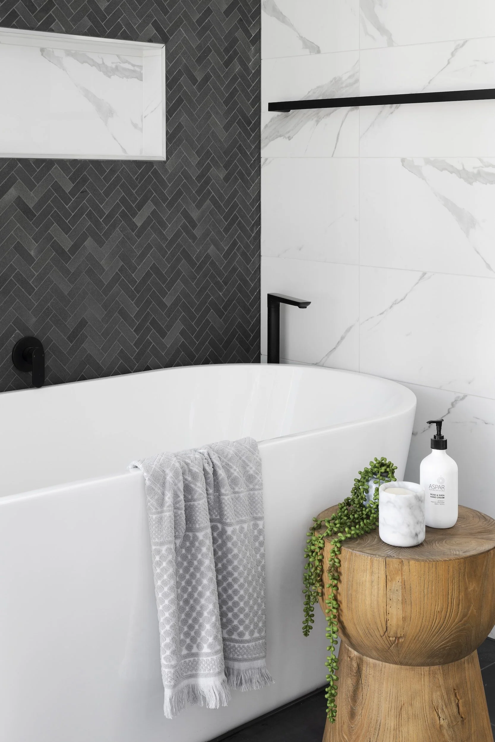 Modern bathroom with a white freestanding tub, black fixtures, a wooden stump side table with a potted plant, hand soap, and a marble container, against a background of white and black marble tiled walls.