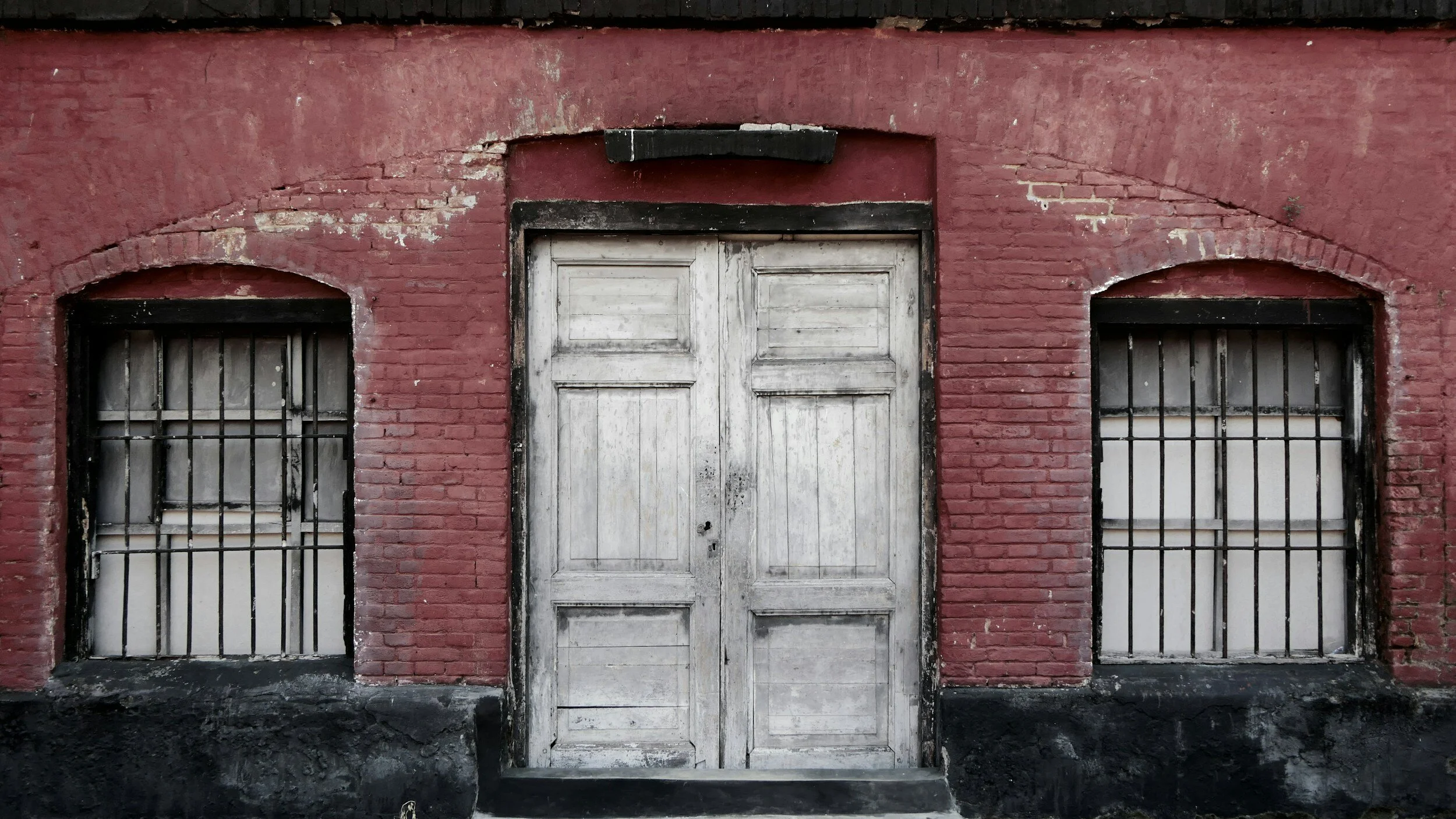 Old, weathered white double door with peeling paint, flanked by two barred windows, set in a red brick building with black accents and worn black foundation.