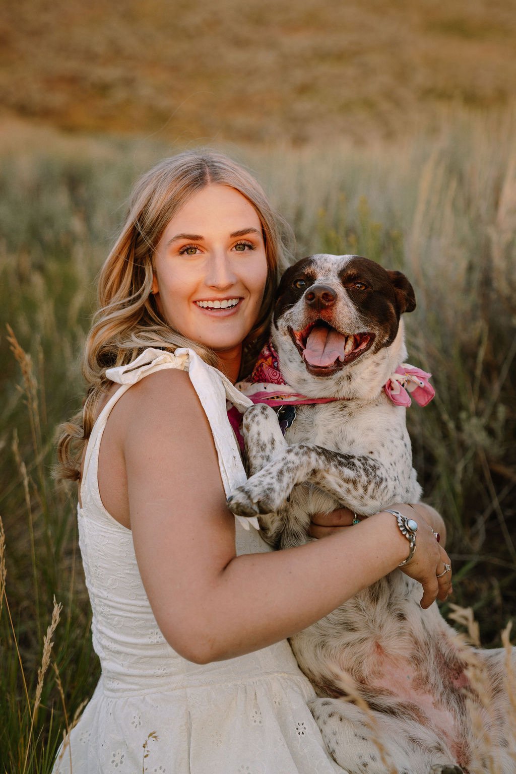 A smiling woman in a white dress holding a happy, excited dog with a bandana around its neck in a grassy field at sunset.