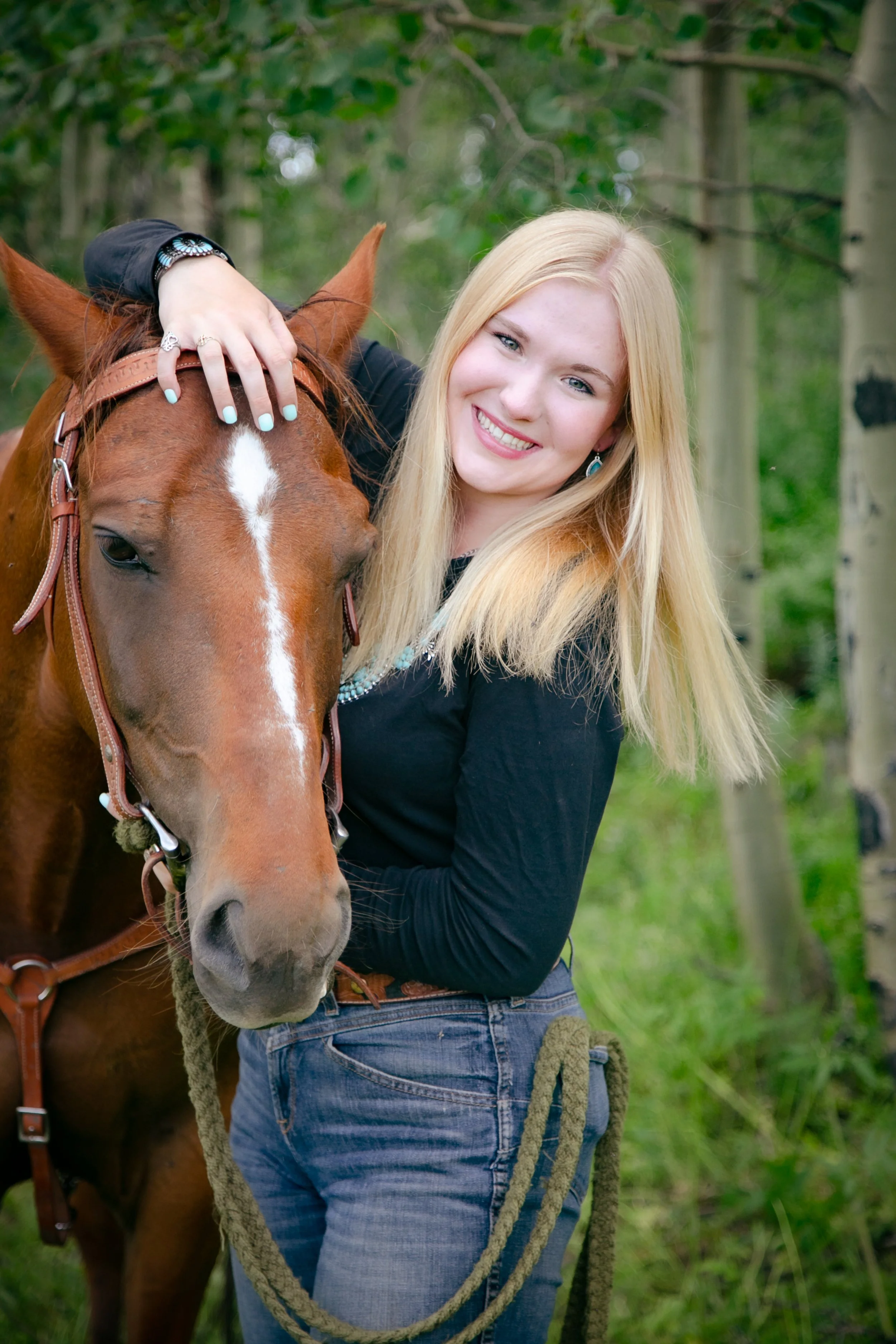A woman with long blonde hair and a black top standing next to a brown horse with a white stripe on its face in a wooded outdoor setting, smiling and touching the horse's head.