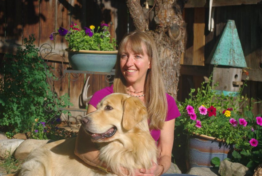 A woman with long blonde hair wearing a purple shirt sitting outdoors with a golden retriever dog, surrounded by colorful flowers and garden decorations.
