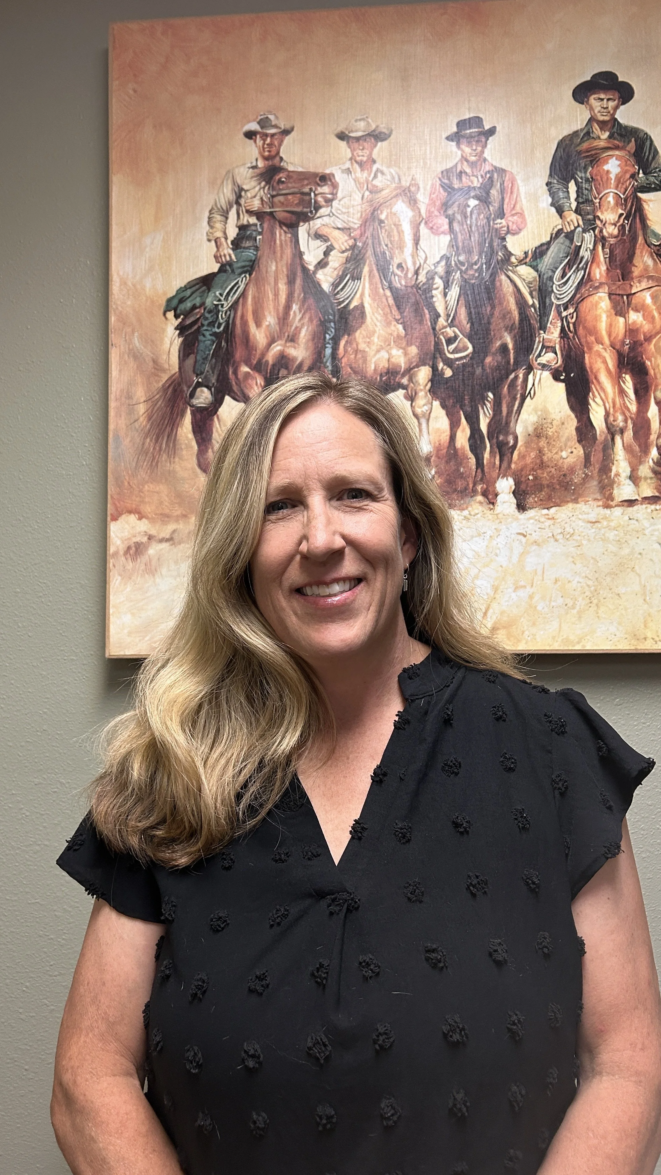 A woman with long blonde hair smiling and wearing a black dress with textured polka dots, standing in front of a painting of four cowboys on horseback.