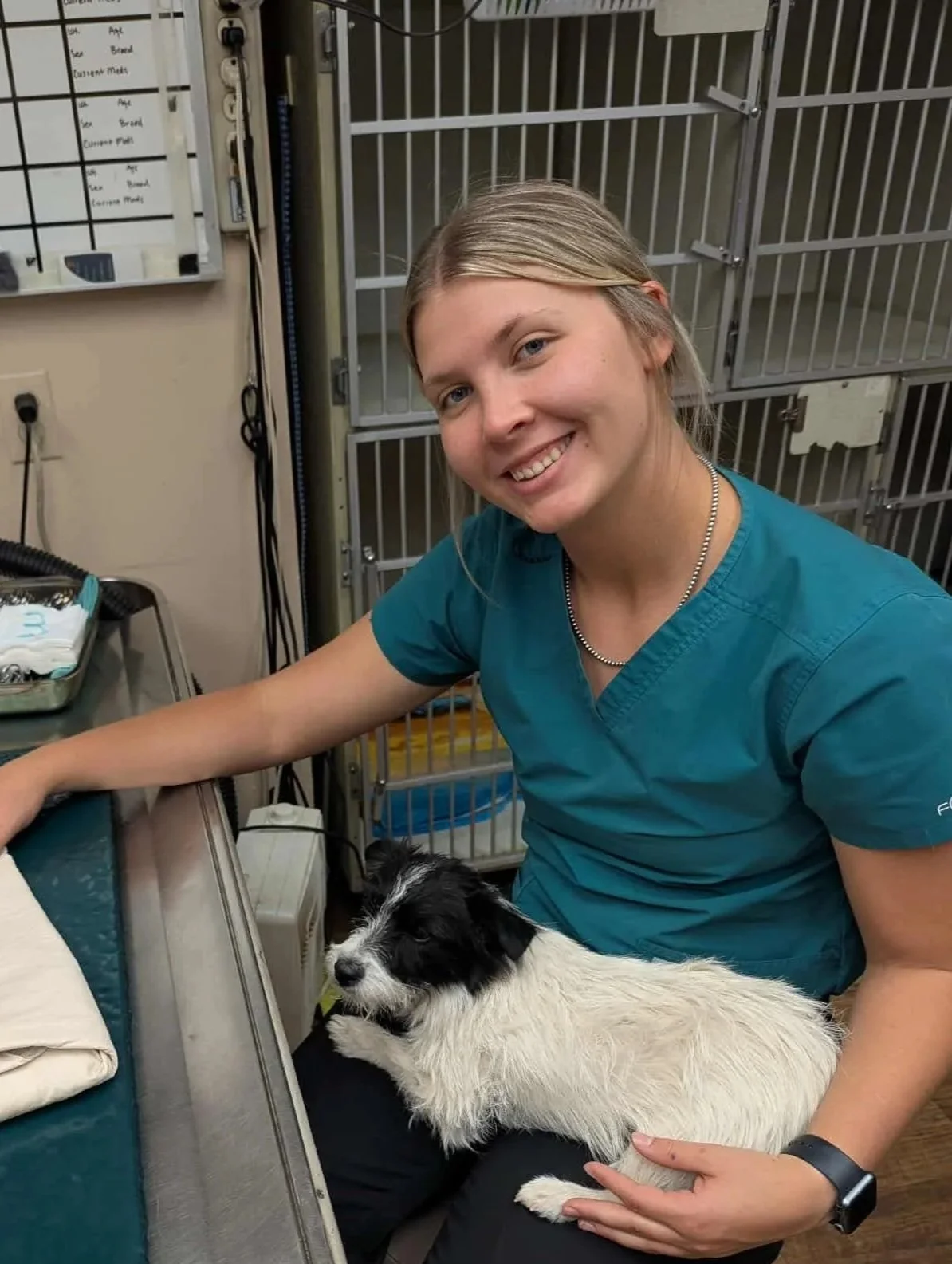A young woman with long blonde hair in a braid, smiling, sitting on a red barn floor, feeding a calf with a bottle. There is a black calf lying beside her, with its head resting on the floor.