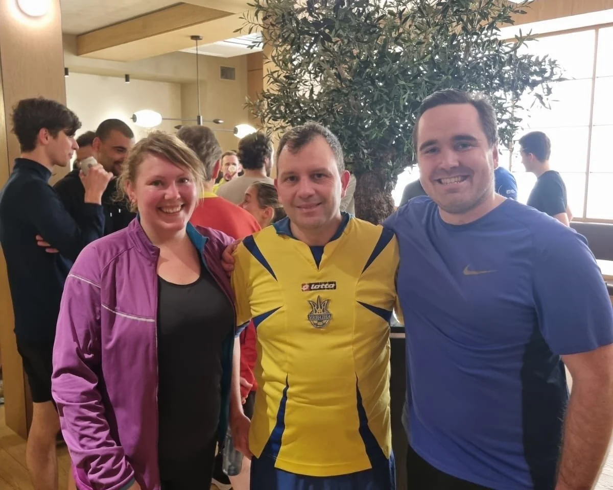 Three people smiling and posing for a photo indoors, with a large tree in the background, among a crowd of people at a social gathering.