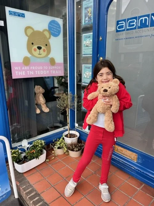 Young girl with dark hair wearing a red jacket and red pants, standing outside a building, hugging a teddy bear, with teddy bears and potted plants around, and a poster with a teddy bear and the words 'We are proud to support the TN Teddy Trail' in the background.