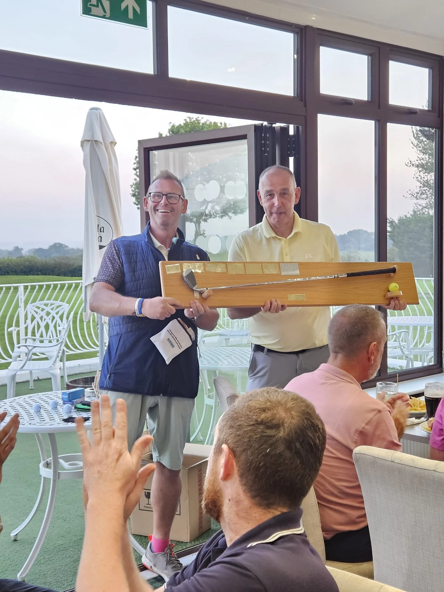 Two men are holding a large wooden golf putter in front of a group at a social gathering indoors with large windows showing an outdoor view of trees and sky. One of the men is smiling, wearing glasses, a dark blue vest, and beige shorts, while the other is in a yellow shirt. Several people are seated at tables, some clapping or raising hands, with drinks and food visible.