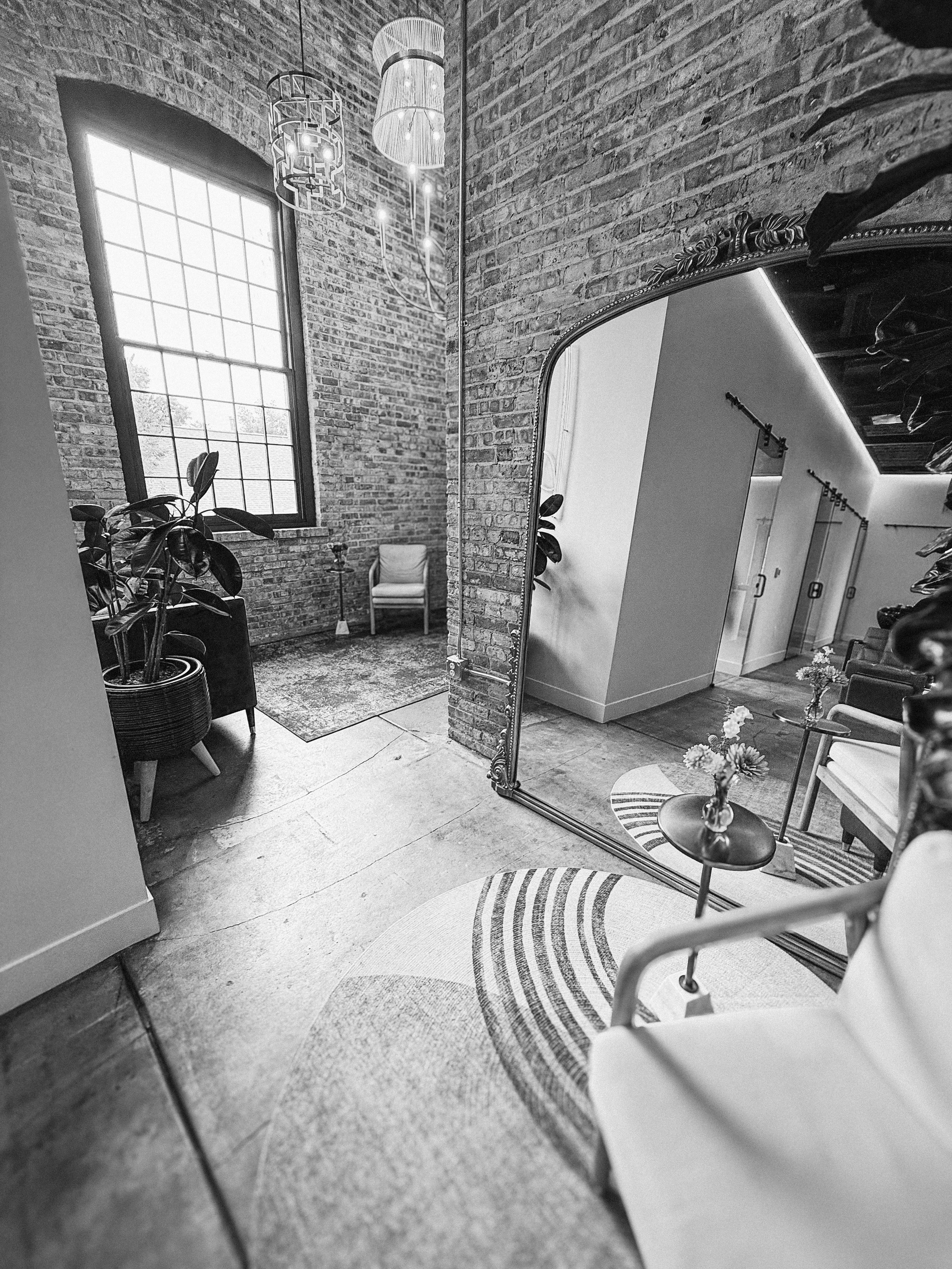 Black and white photo of an interior space with a large brick wall, tall window, and various furniture including a mirror, chairs, a small table with a vase of flowers, potted plants, and modern lighting fixtures.