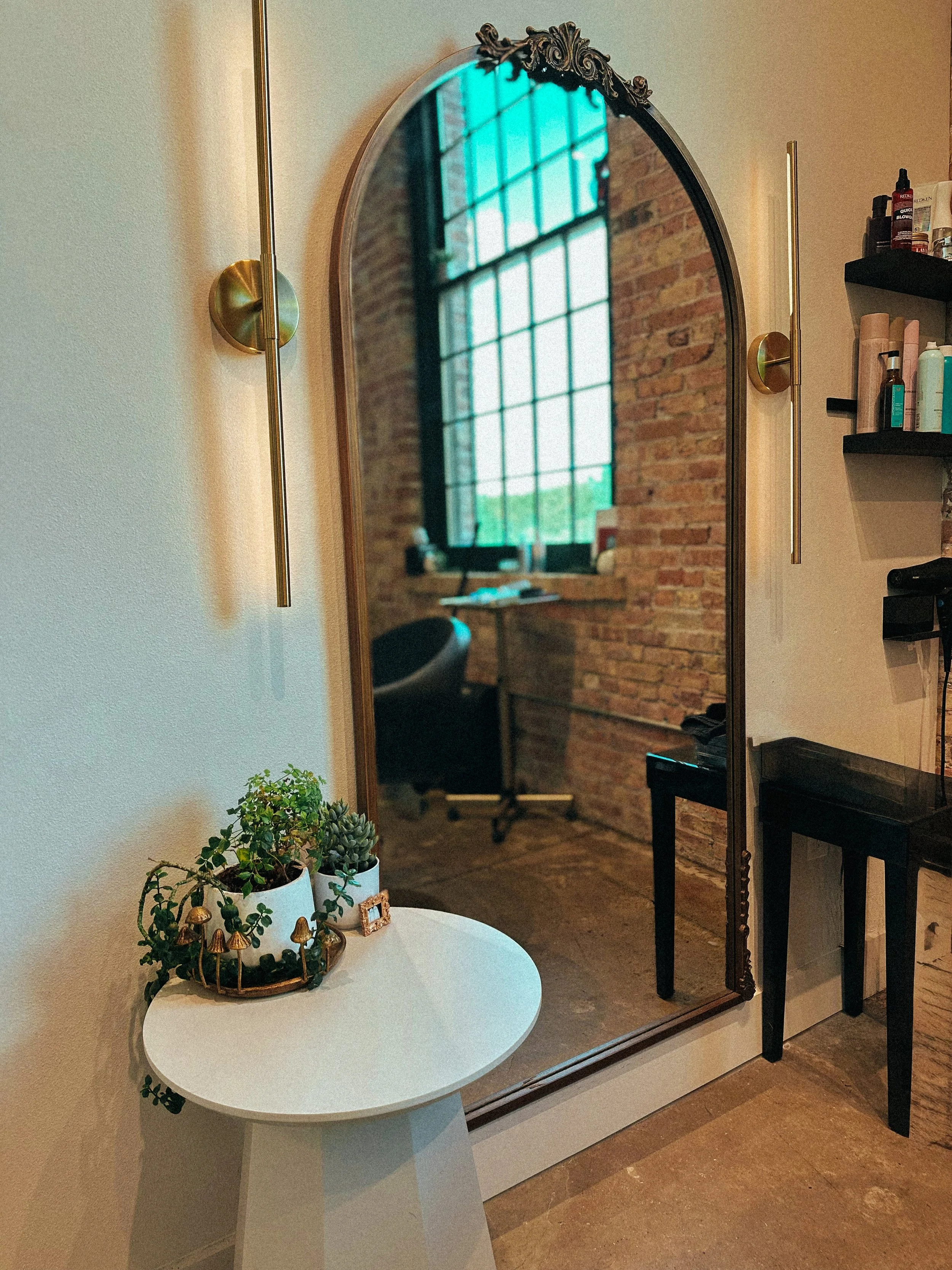 Interior of a room with a decorative mirror, black shelf with various skincare products, a table with plants, and a brick wall with large windows.