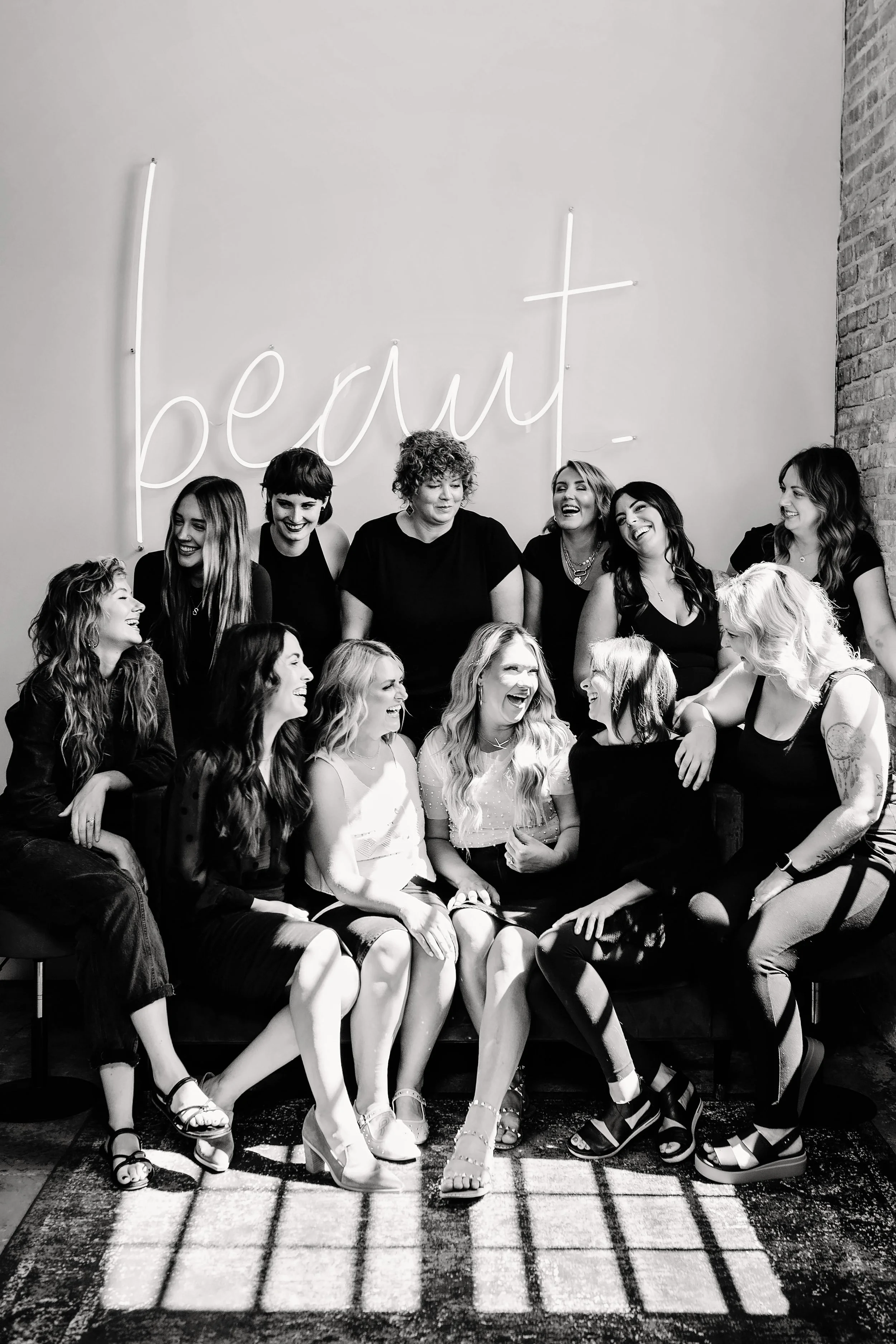 Black and white photo of a group of women sitting and standing together, smiling, in front of a wall with a neon sign that says 'be your' and a brick wall on the right.