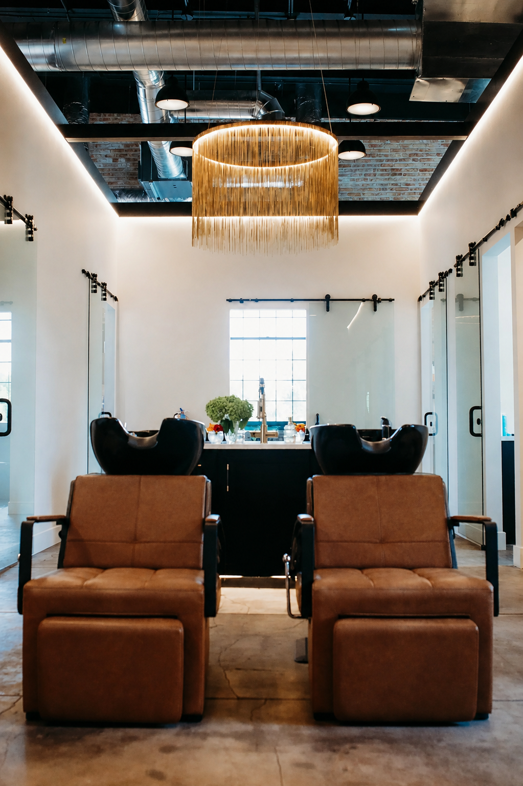 A modern hair salon with two styling chairs facing mirror and wash stations. Brown leather seats in the foreground, black shampoo bowls in the background, and a large, circular chandelier with hanging fringe overhead. Bright natural light from a window at the back.