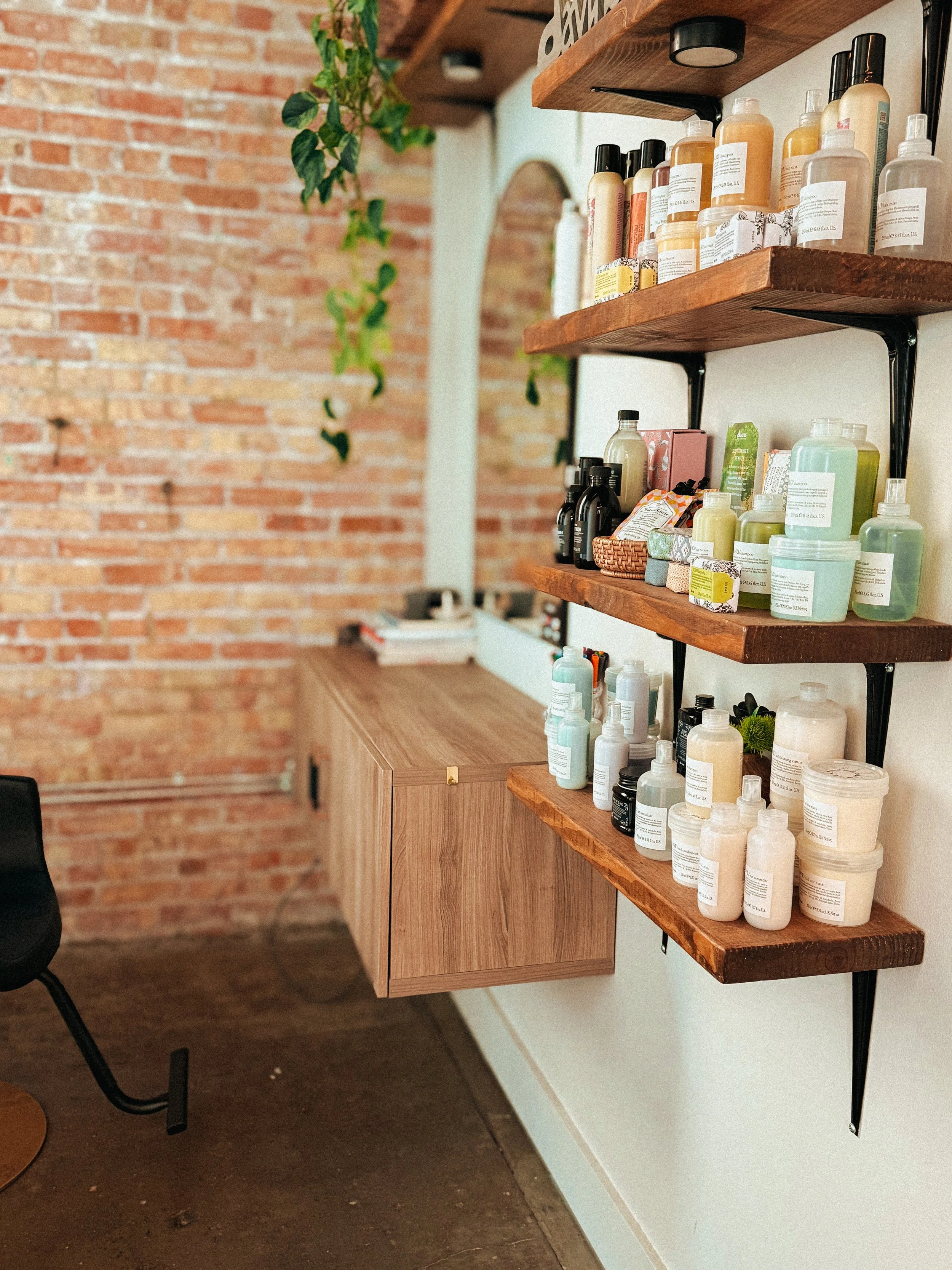 Wooden shelves mounted on a wall with various bottles of skincare or personal care products, in a room with a brick wall and wooden furniture.