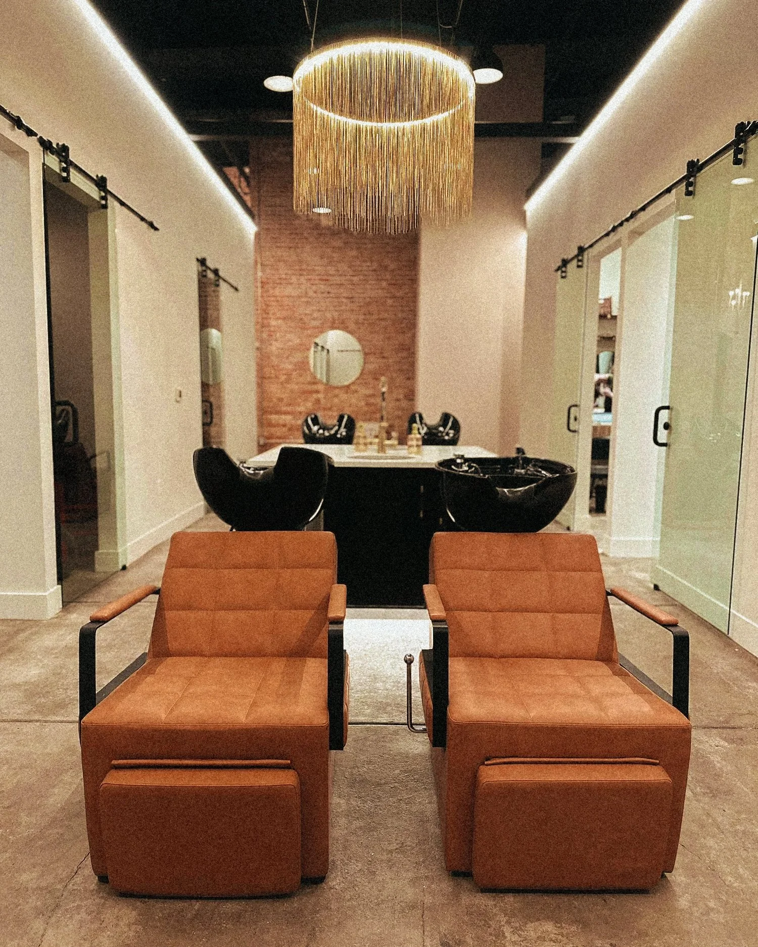 A waiting area or lobby with two brown armchairs in the foreground and a black table with drinks behind them. In the background, a dining or meeting area with a white table, four black chairs, and a decorative circular light fixture hanging from the 
