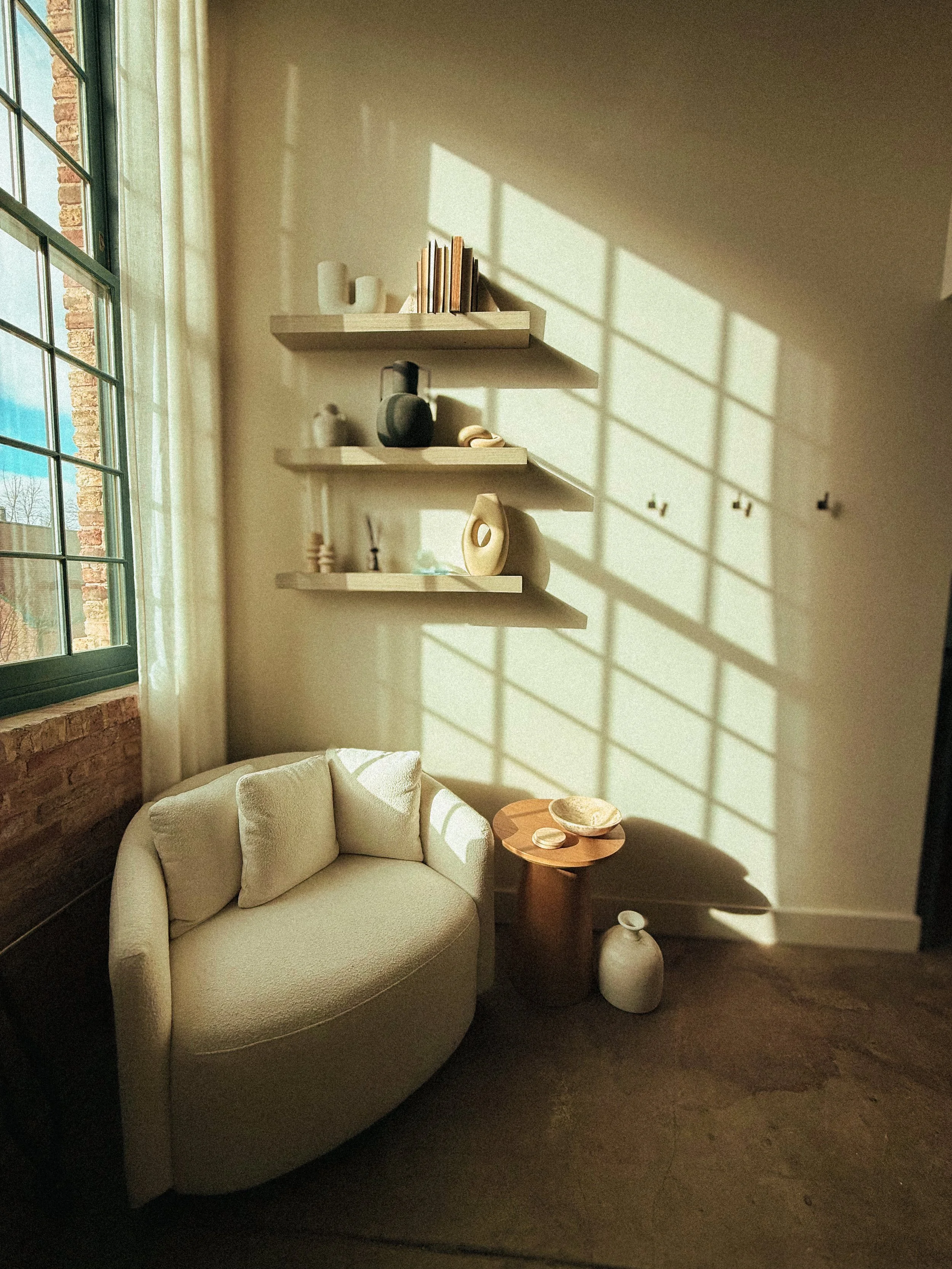 A cozy living room corner with a cream-colored, rounded sofa with pillows, a small wooden side table holding decorative bowls, and white vases on wall shelves. Sunlight creates geometric shadows on the wall.