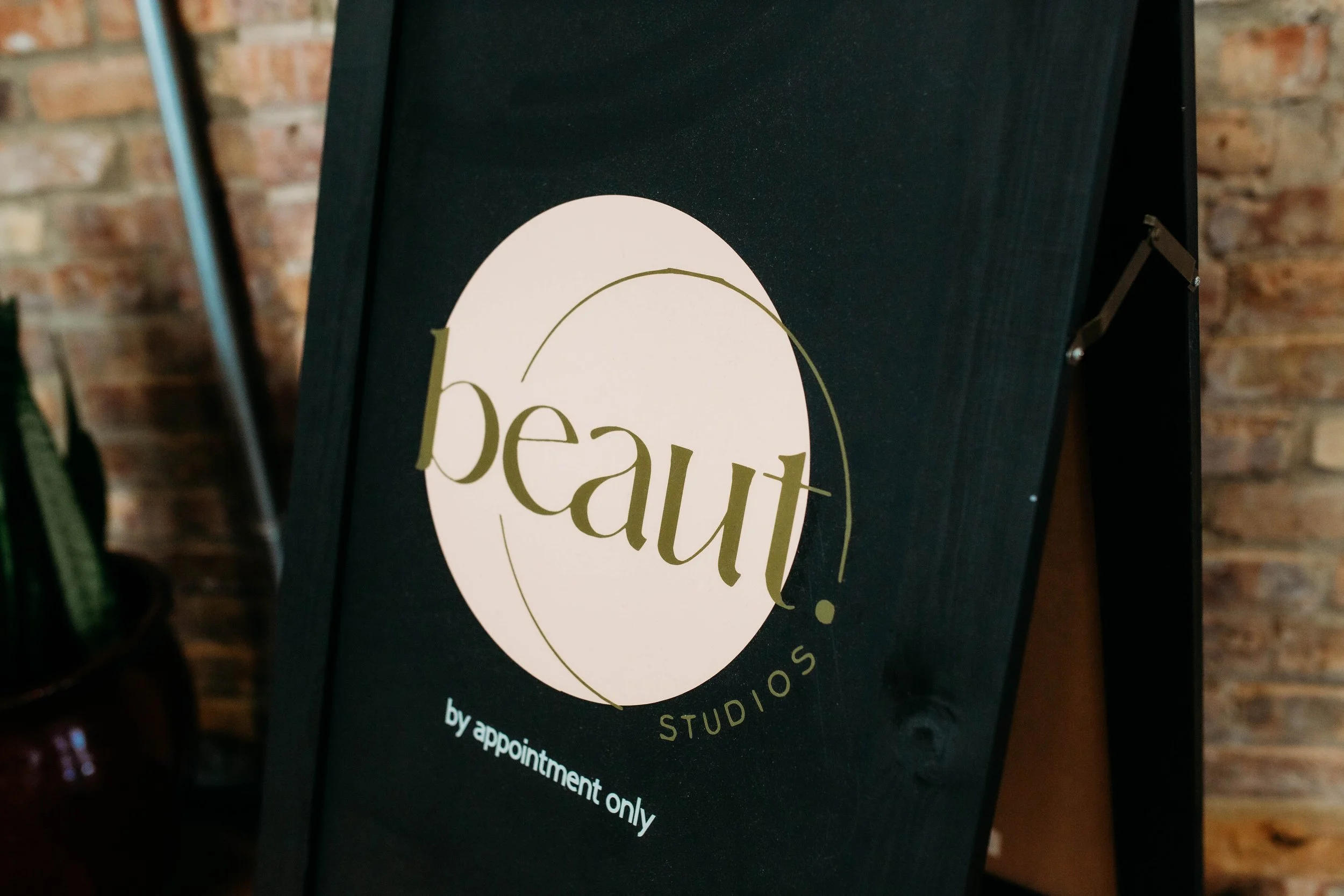 A black sign with a white oval shape and gold text that reads, 'Beauty Studio'. The sign has smaller text below that says, 'by appointment only'. In the background, there is an exposed brick wall.