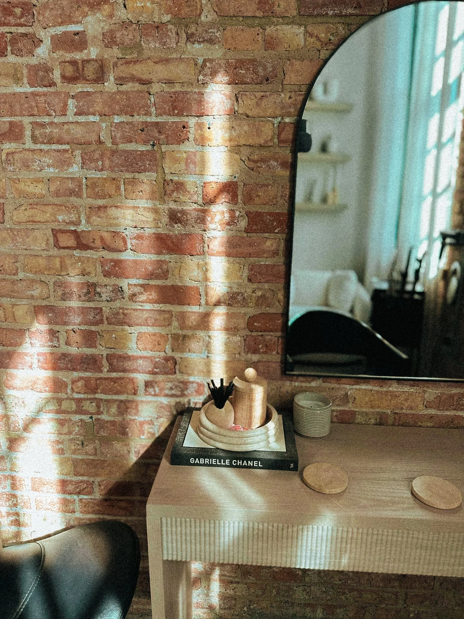 A corner of a room with a brick wall, a small beige table, and a mirror. On the table, there is a black book titled 'GABRIELLE CHANEL', and a wooden tray holding small objects including a beige container with black straws, a small wooden figure, and 