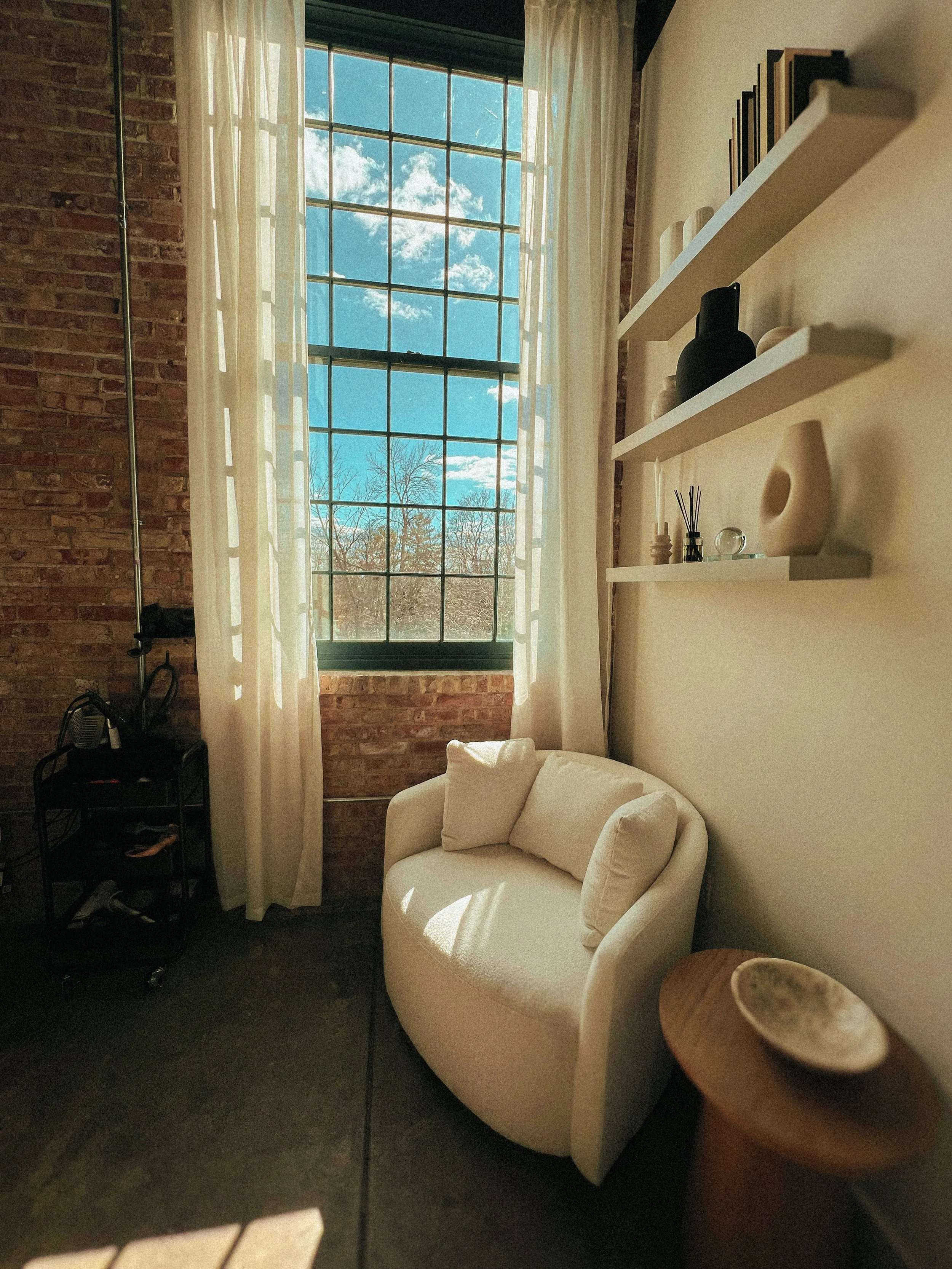 A cozy corner of a room with a large window showing blue sky and trees, a white sofa, floating shelves with books and decor, and a brick wall adjacent to the window.