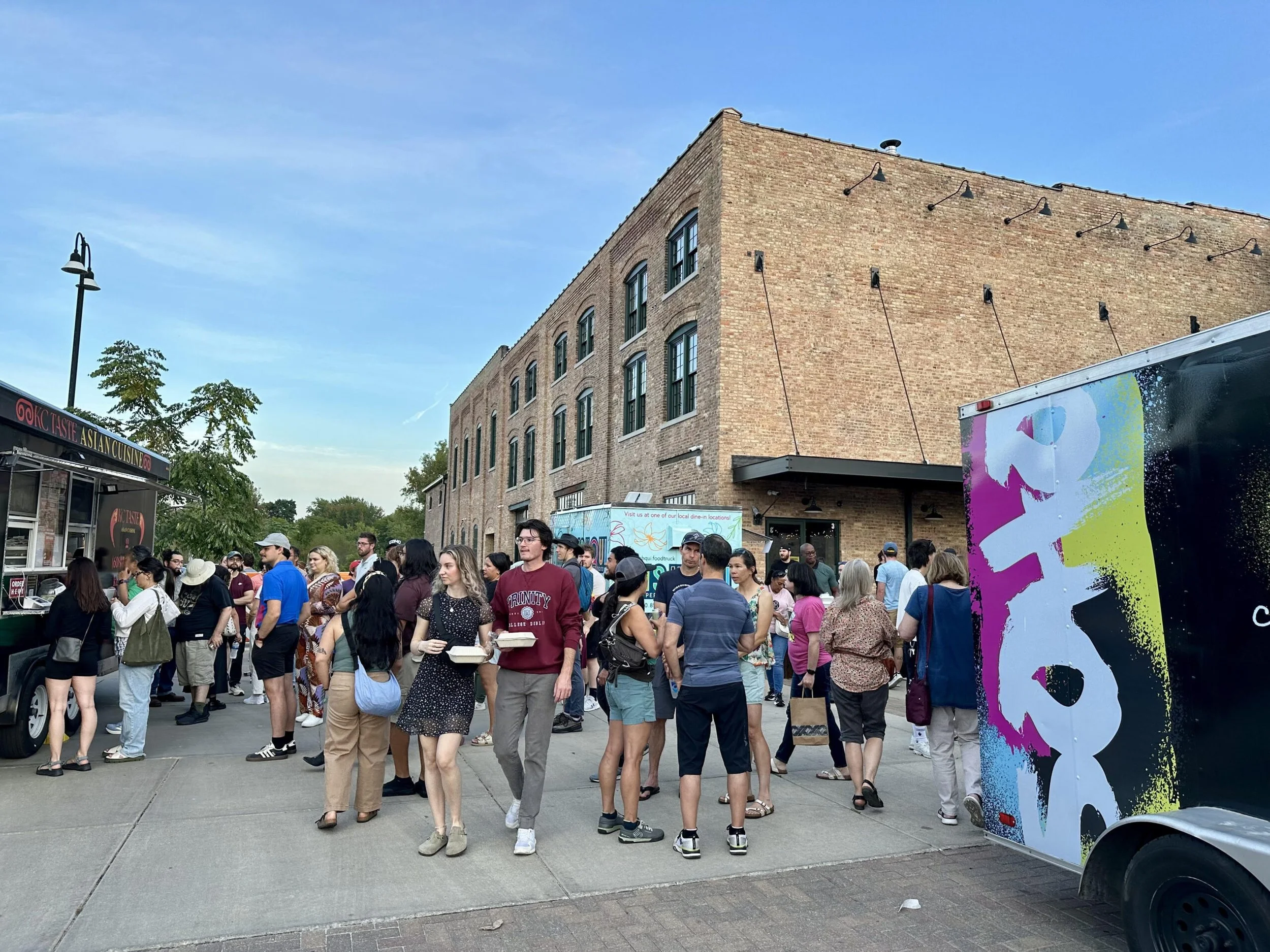 Crowd of people standing outside near a brick building with outdoor lighting fixtures and a colorful truck with abstract art.