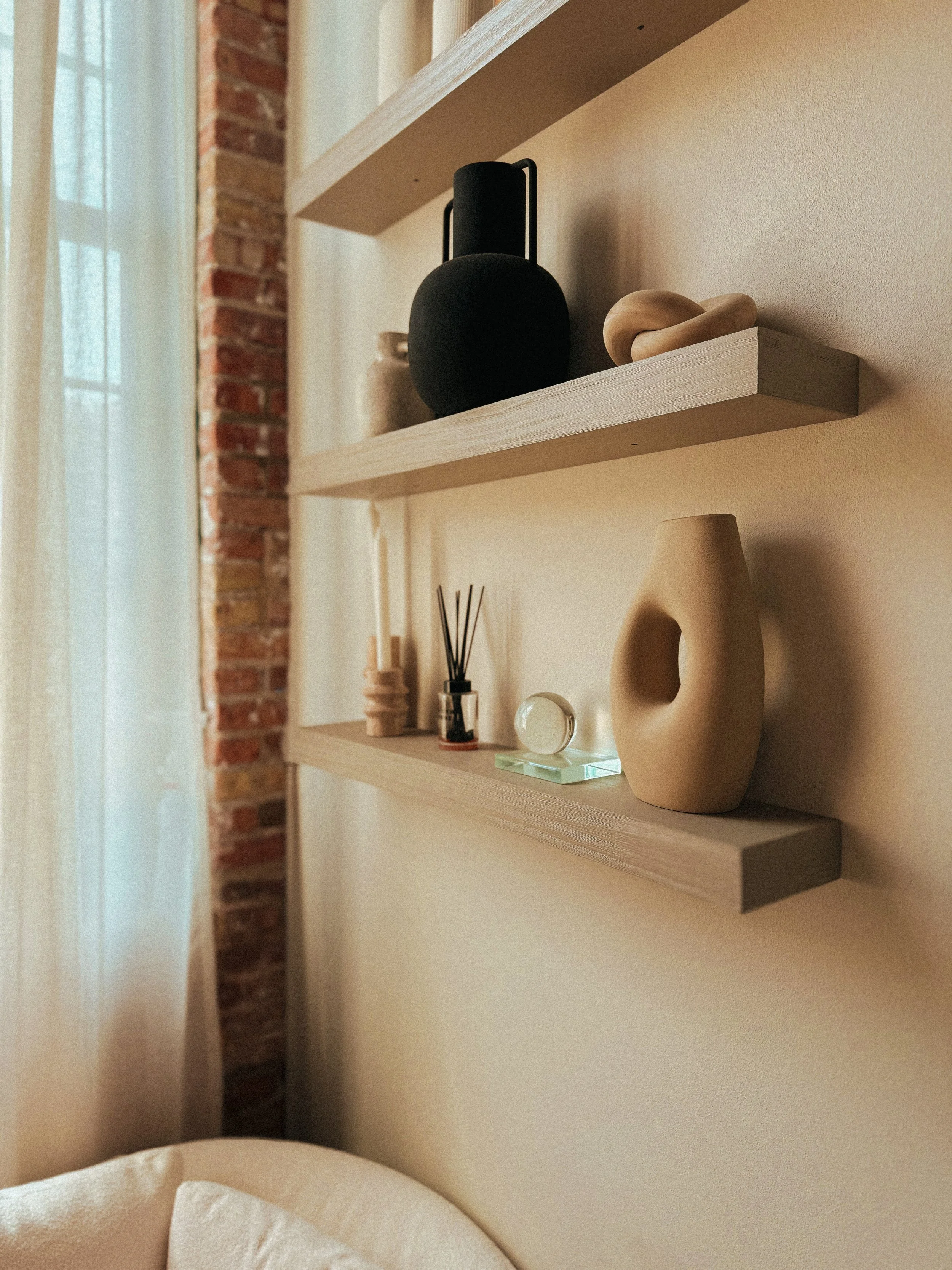 A close-up image of two wooden shelves mounted on a beige wall, decorated with various objects including a black textured speaker, ceramic vases, a reed diffuser, and a spherical object, with part of a bed and a brick wall with a window curtain visib