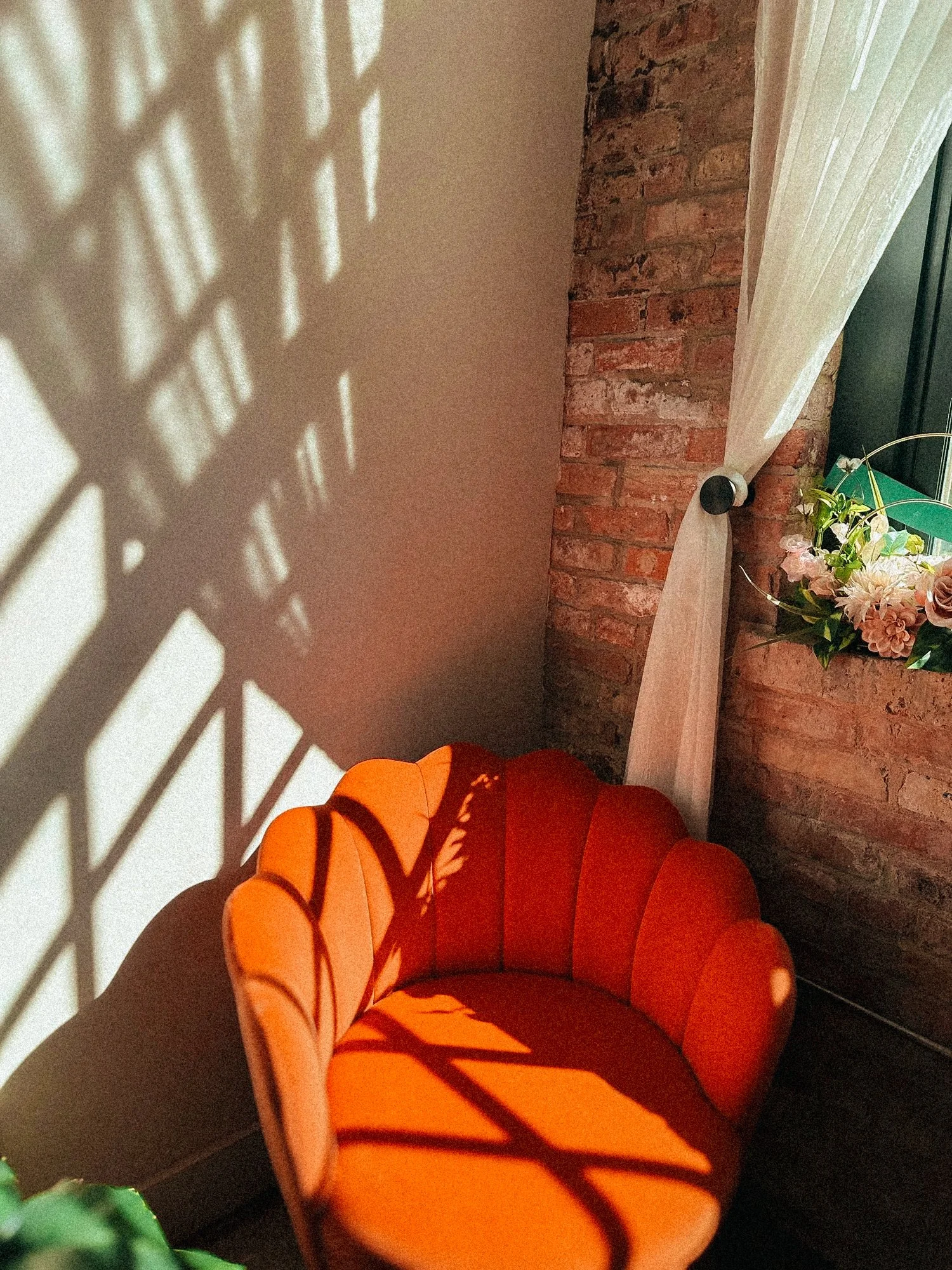 A red velvet, barrel-shaped armchair with a velvety texture, situated in a corner with exposed brick wall on one side and a white wall on the other. Shadows cast by the window create a criss-cross pattern on the white wall. There is a floral arrangem