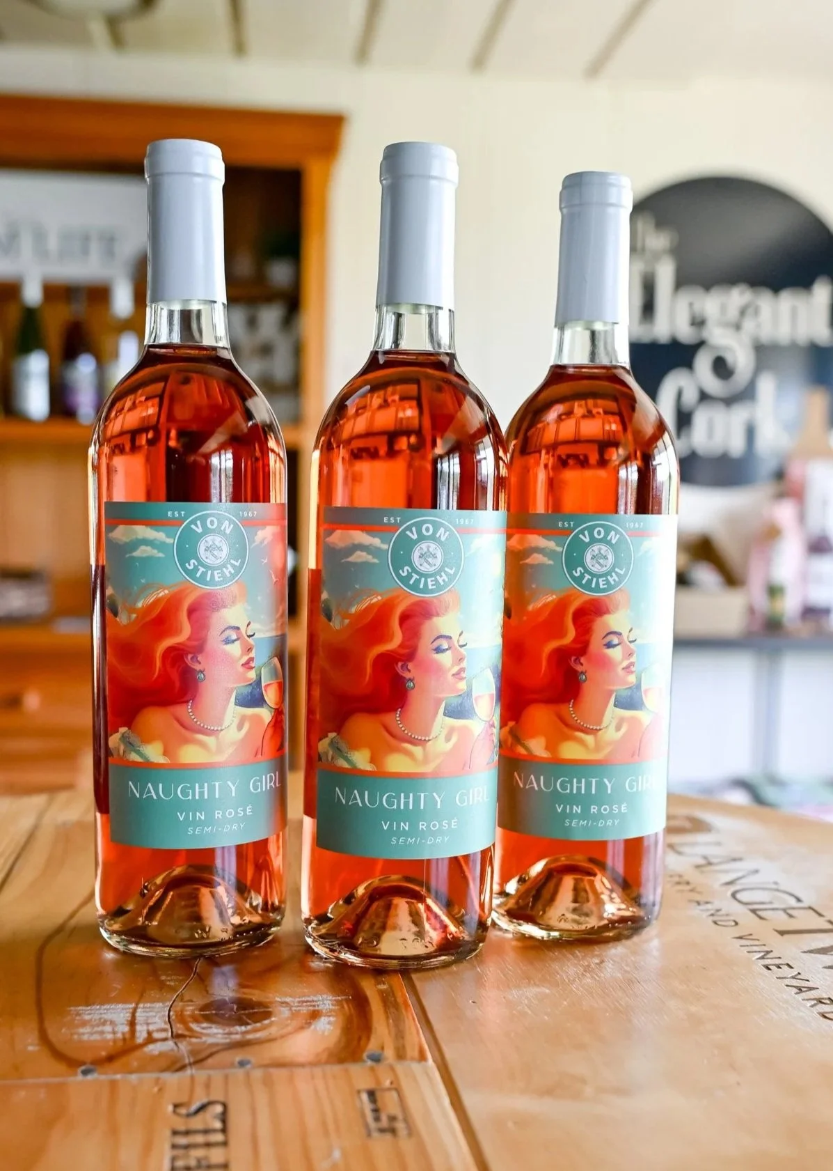 Three bottles of NV Naughty Girl Vin Rosé semi-dry wine with colorful vintage artwork of a glamorous woman with red hair, pearls, and wine glass.