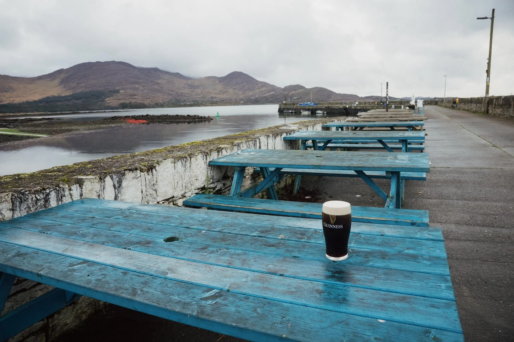 A fresh pint of Guinness sitting on a weathered blue picnic table at the water's edge in Kilmakilloge, looking out over a quiet bay toward the misty mountains of the Beara Peninsula.