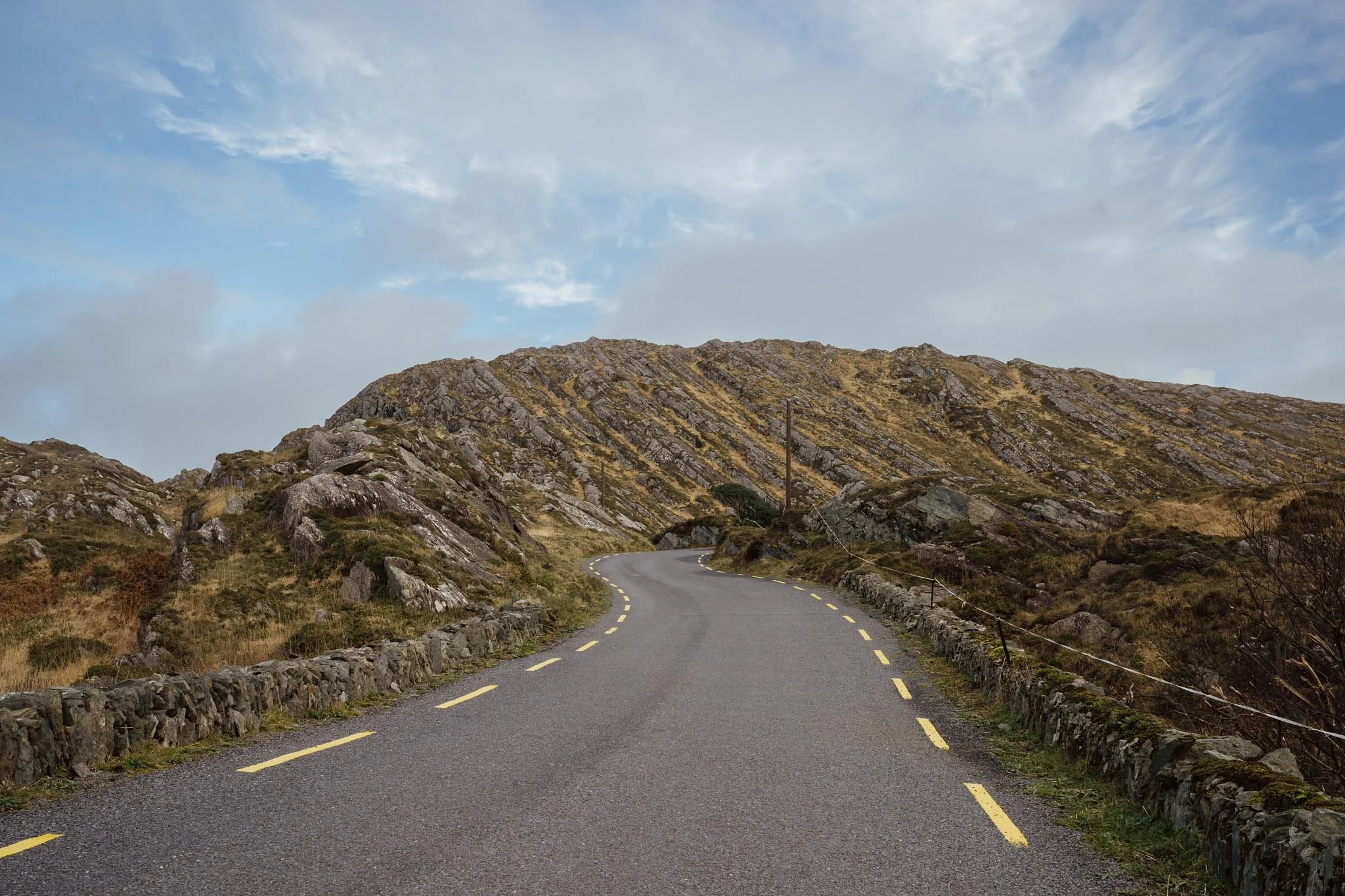 A view looking up a steep, curving road on a rocky hillside in West Cork, framed by low stone walls and jagged, sunlit terrain.
