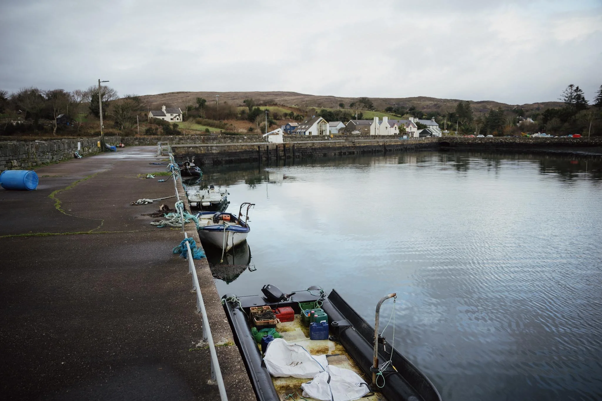 A wide, moody view across the still waters of Kilmakilloge Bay toward a silhouette of layered mountains, with a stone pier and a small green navigation marker visible in the foreground.