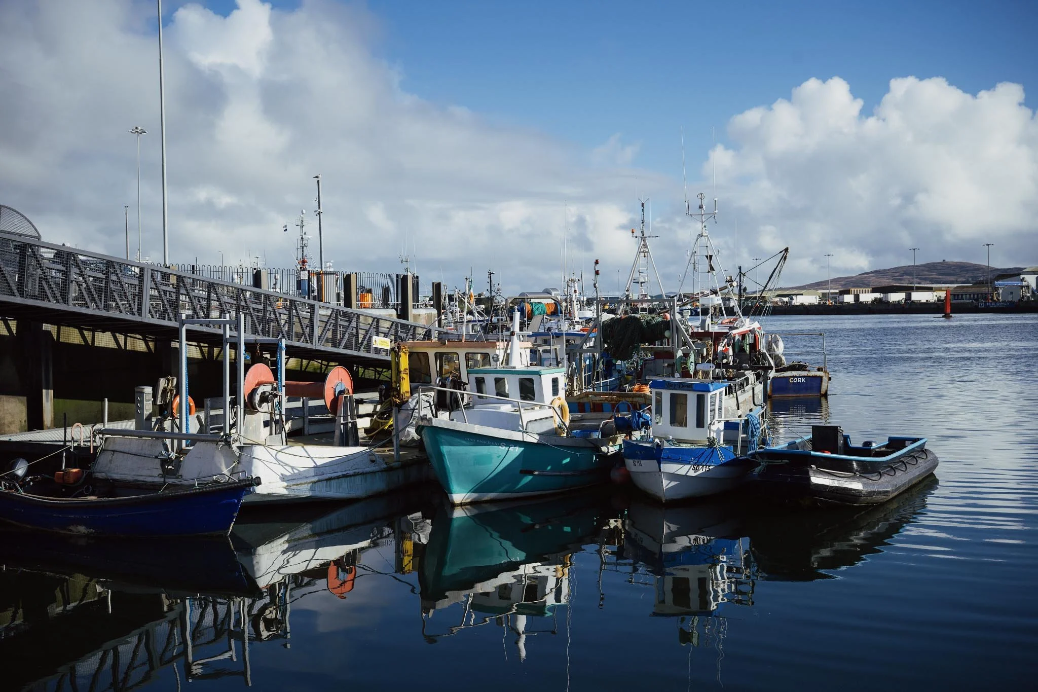 A group of colorful commercial fishing boats moored at a metal pier in Castletownbere harbor, County Cork, with their reflections shimmering in the calm blue water under a bright, cloudy sky.