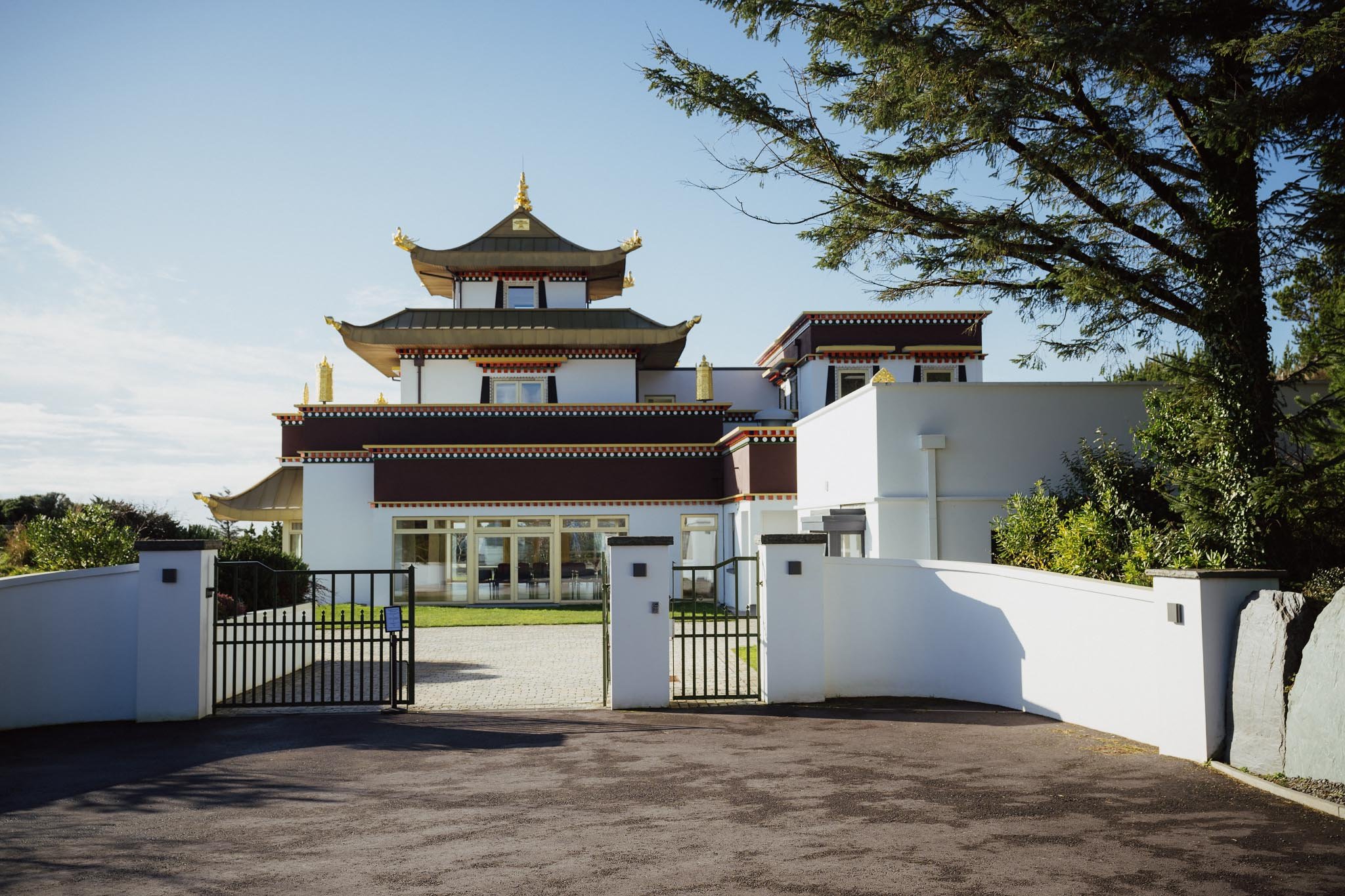 The front entrance of the Dzogchen Beara temple building, featuring traditional white walls, maroon accents, and tiered golden roofs under a bright sun.