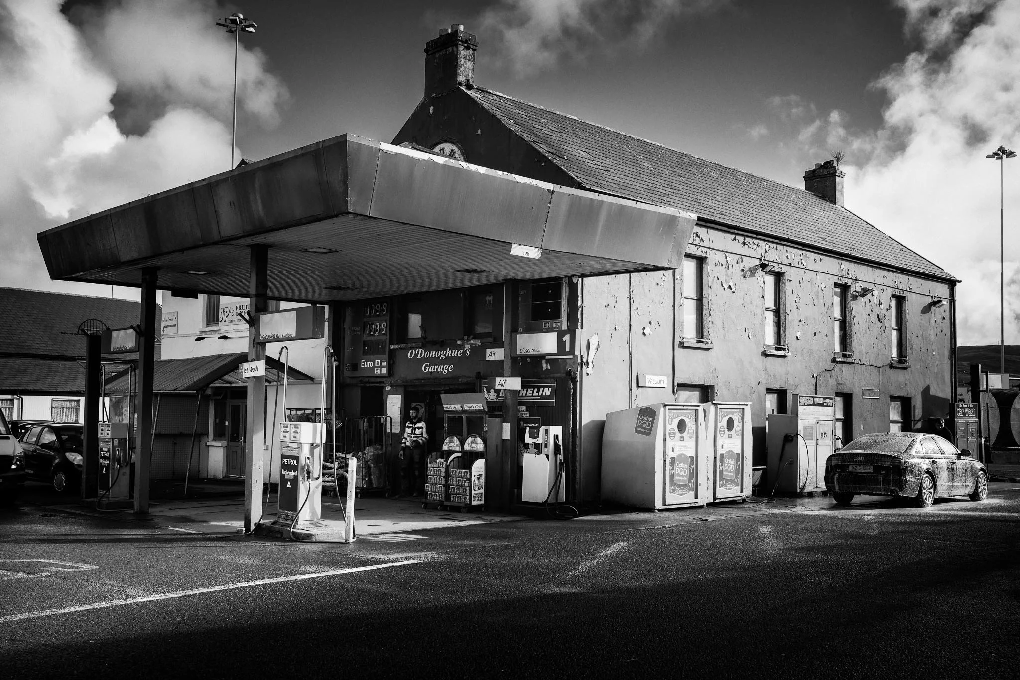 A dramatic black and white photograph of O'Donoghue's Garage in Castletownbere. The image shows a large weathered canopy over old petrol pumps, a textured stone building with peeling paint, and a car being washed in the foreground.