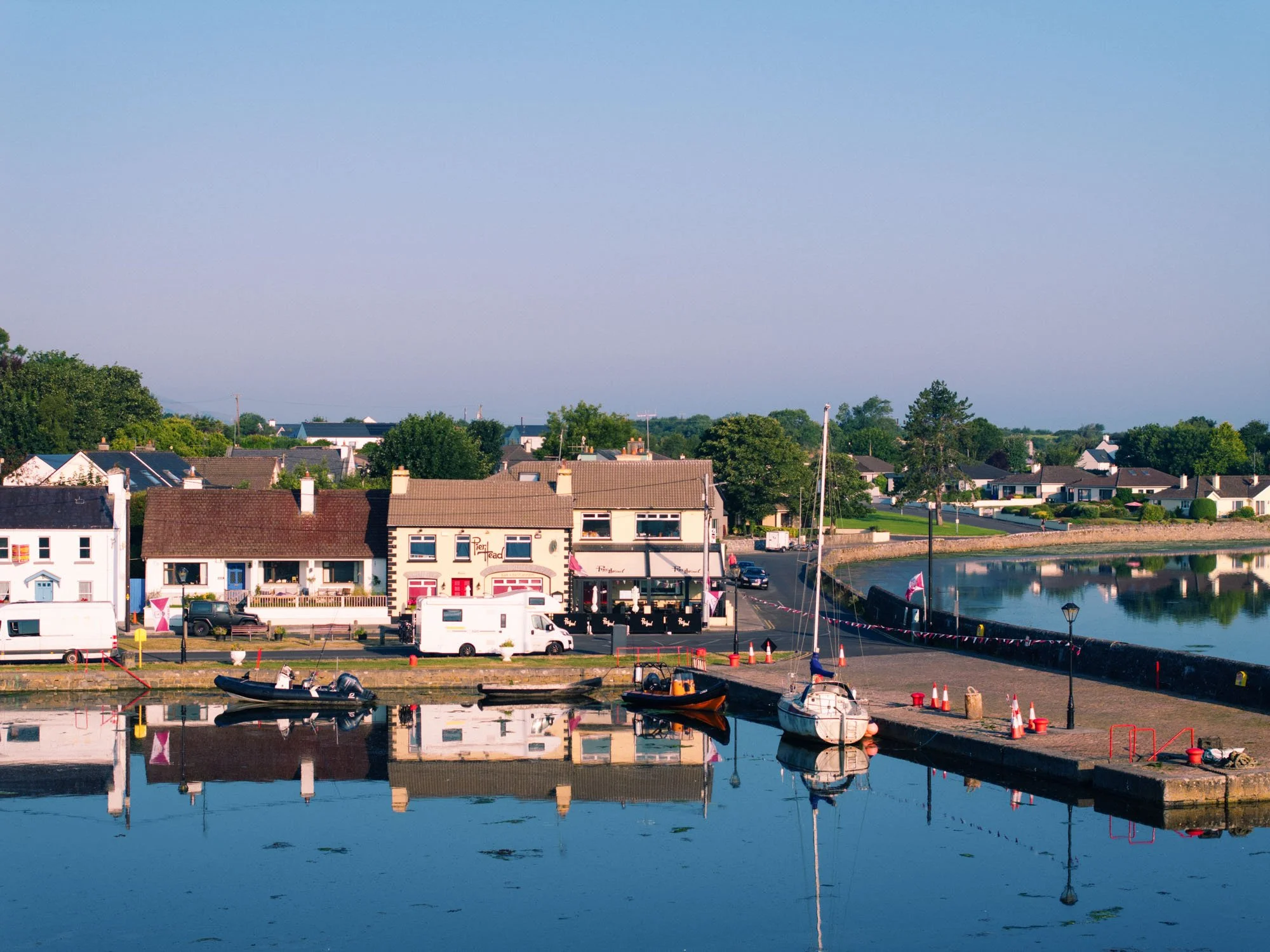Aerial view of Kinvara harbour beside the Pier Head pub, with small boats and a sailboat moored along the quay, a campervan parked on the waterfront, and still water reflecting the buildings under a clear evening sky