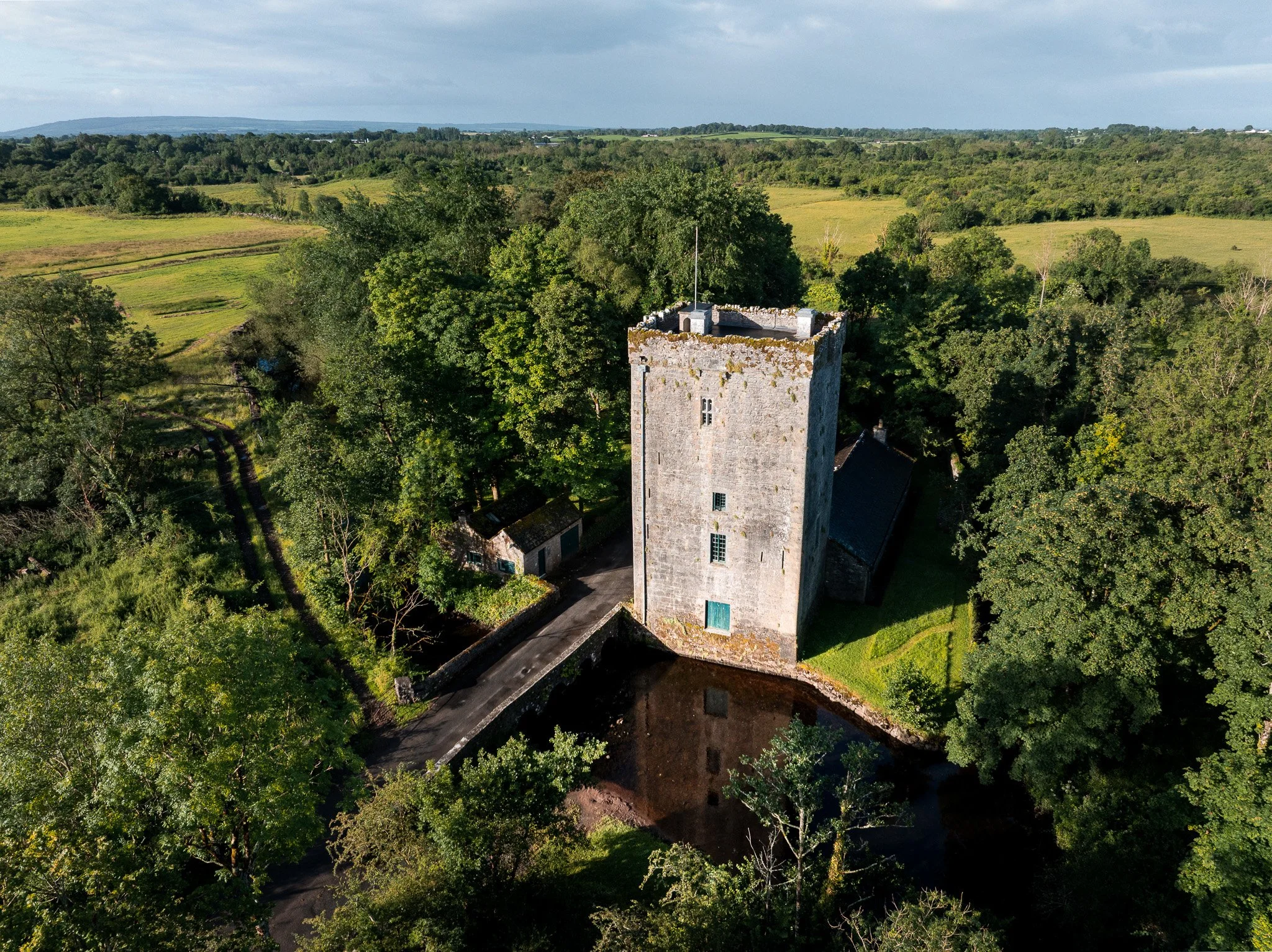 Aerial view of Thoor Ballylee, the medieval Yeats tower house near Gort, County Galway, with its riverside setting, adjoining cottage, and green farmland stretching to distant hills.